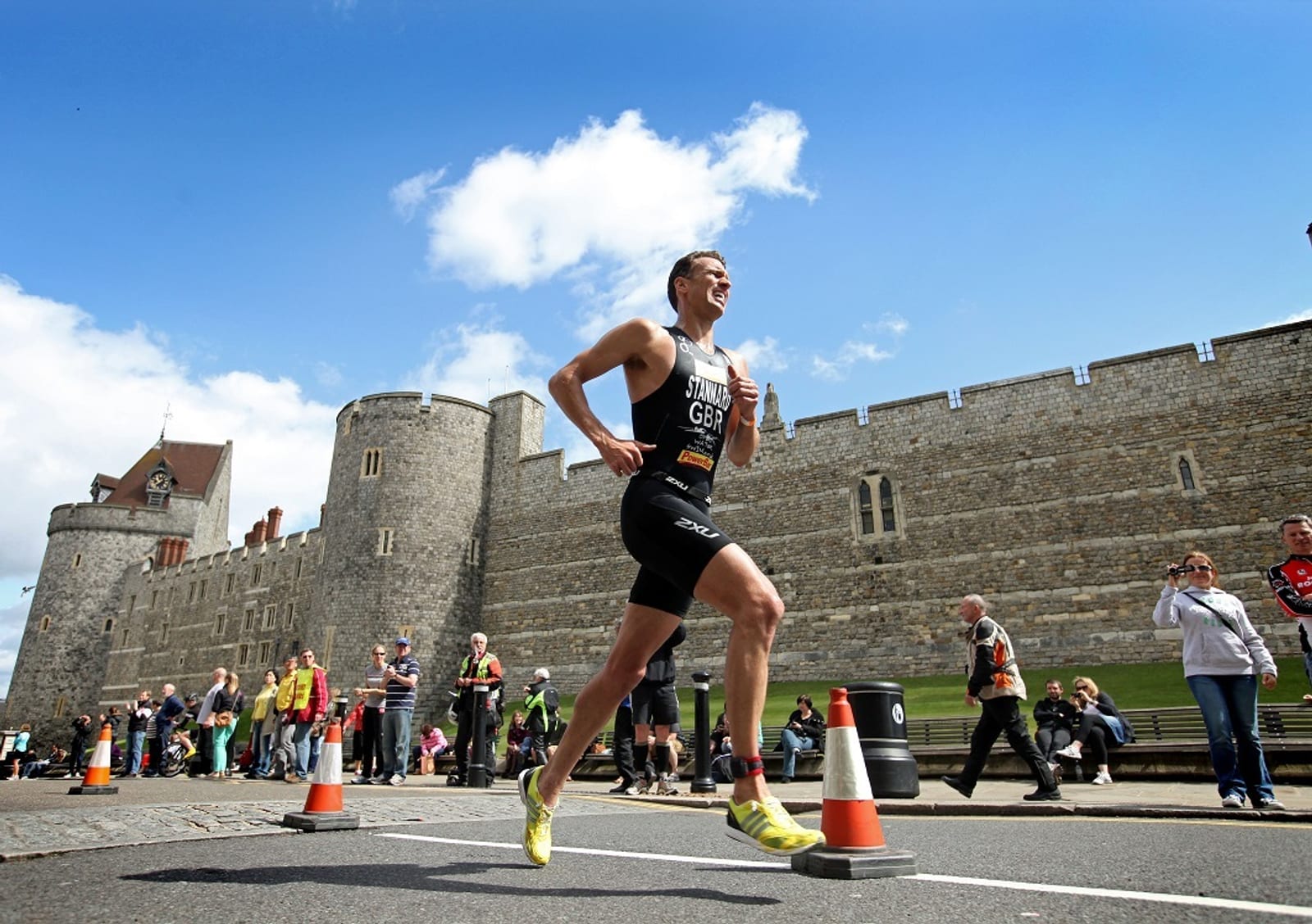 A triathlete runs past Windsor Castle in a race. The athlete, wearing a black suit with "GBR" printed on it, is surrounded by cheering spectators. An orange traffic cone marks the race path. The sky is bright blue with scattered clouds.