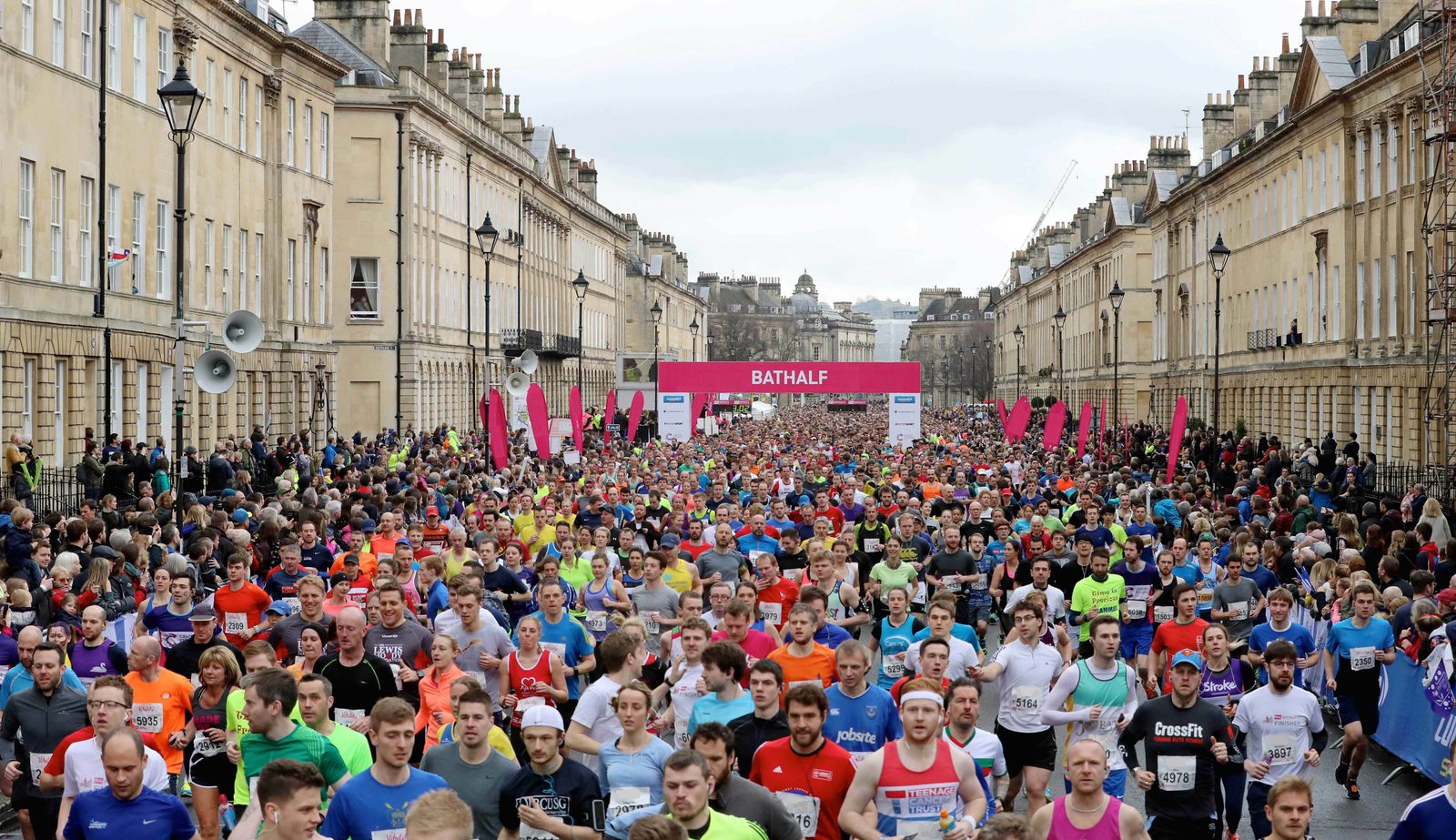A large crowd of runners participates in a marathon event down a street lined with historic buildings. The runners wear colorful athletic gear and bib numbers. Spectators watch from the sidelines, and a pink banner with "BATHALF" is visible in the background.