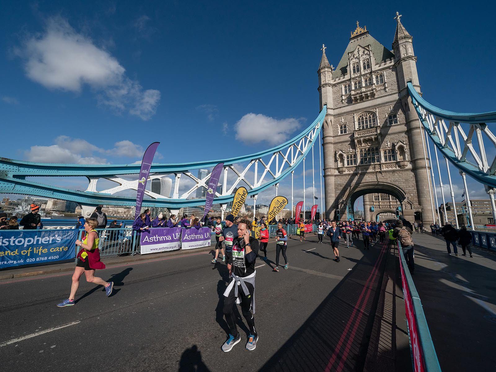 A group of marathon runners crossing the iconic Tower Bridge in London on a sunny day. The participants run past colorful banners and cheering crowds. The bridge's towering structure and blue sky with scattered clouds form the backdrop.