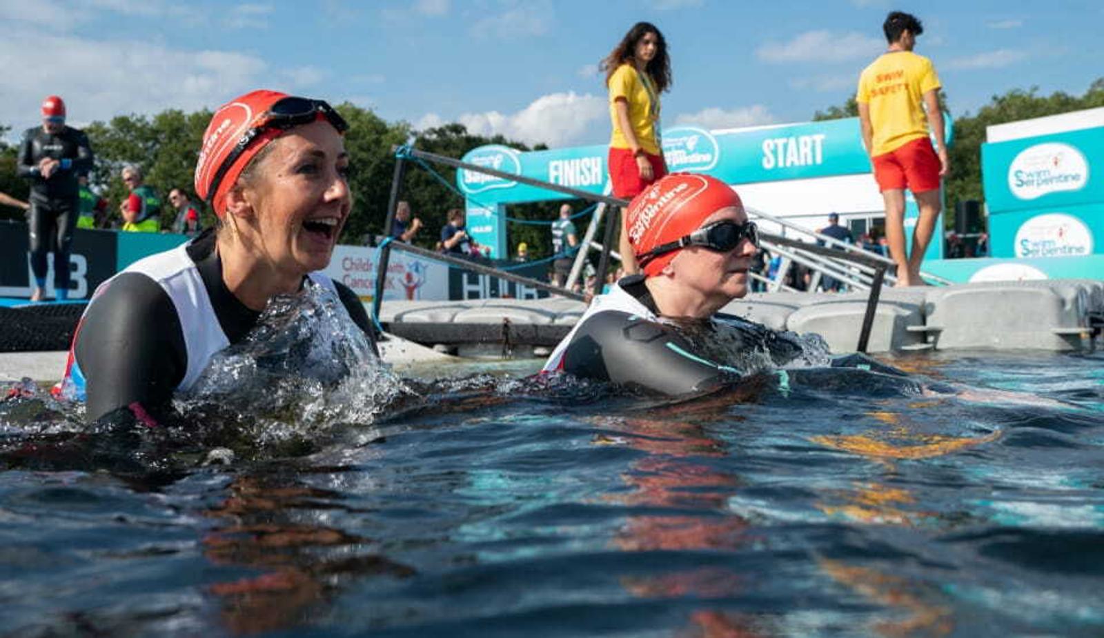 Two swimmers in wetsuits and red swim caps emerge from the water, smiling and looking content at a triathlon event. Others, including participants and supporters in yellow shirts, stand in the background near the finish and start signs. The sky is clear and sunny.