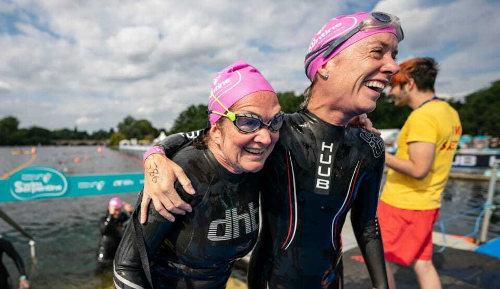 Two swimmers in pink swim caps and black wetsuits stand arm-in-arm at the edge of a body of water, smiling happily. They have just finished a swim event. A person in a yellow shirt and red shorts stands in the background, with other swimmers coming out of the water.