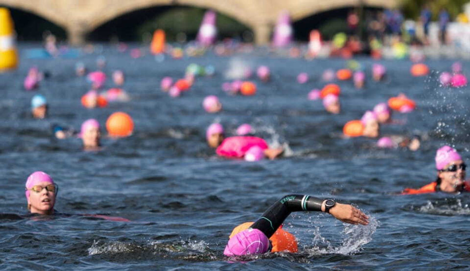 A large group of swimmers in colorful swim caps participate in an open water race. Swimmers are spread across the water, with some in mid-stroke. The background shows a bridge and buoys marking the course. The atmosphere appears lively and competitive.
