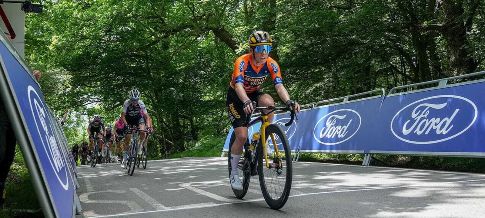 A cyclist in an orange and blue jersey leads a group of riders uphill during a race. The road is lined with metal barriers displaying the Ford logo. Lush green trees and foliage surround the scene on a sunny day.