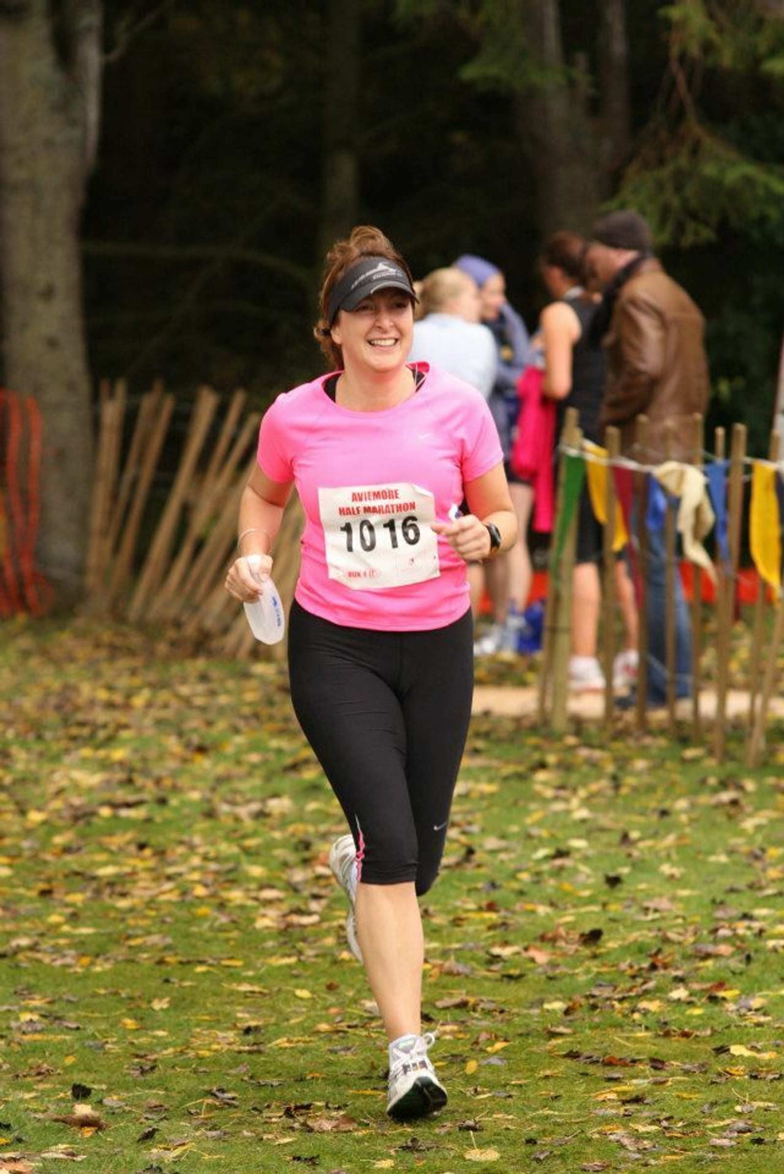 A woman in a bright pink shirt and black leggings, wearing a visor and sunglasses, is running outdoors on a grassy path. She has a race bib numbered 1016 pinned to her shirt and is smiling. Other participants and onlookers are in the background, with autumn leaves on the ground.