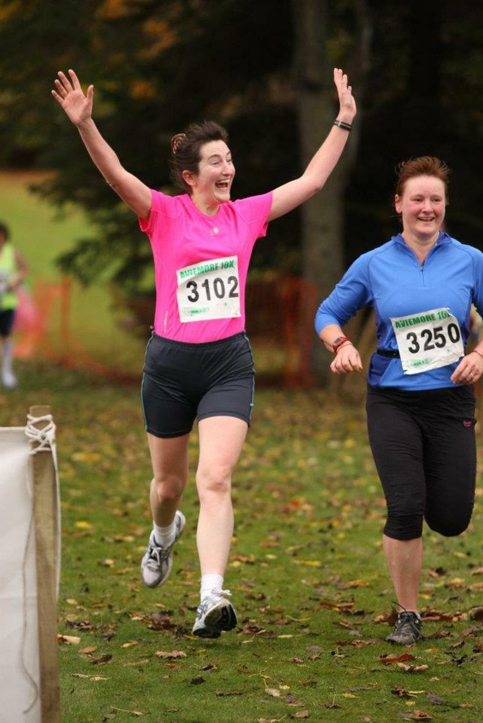 Two women are running a race, both with bib numbers: one (3102) wearing a pink shirt and black shorts, and the other (3250) in a blue shirt and black pants. The woman in the pink shirt, with her arms raised and a joyful expression, looks excited as they run outdoors.
