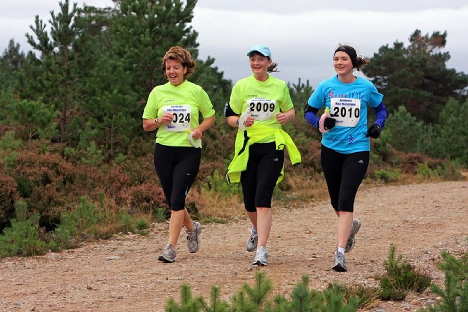 Three women running on a dirt trail surrounded by greenery. They are all wearing athletic gear and race bibs with numbers 324, 2009, and 2010. The woman on the left and center are in bright green shirts, while the woman on the right is in a blue shirt.