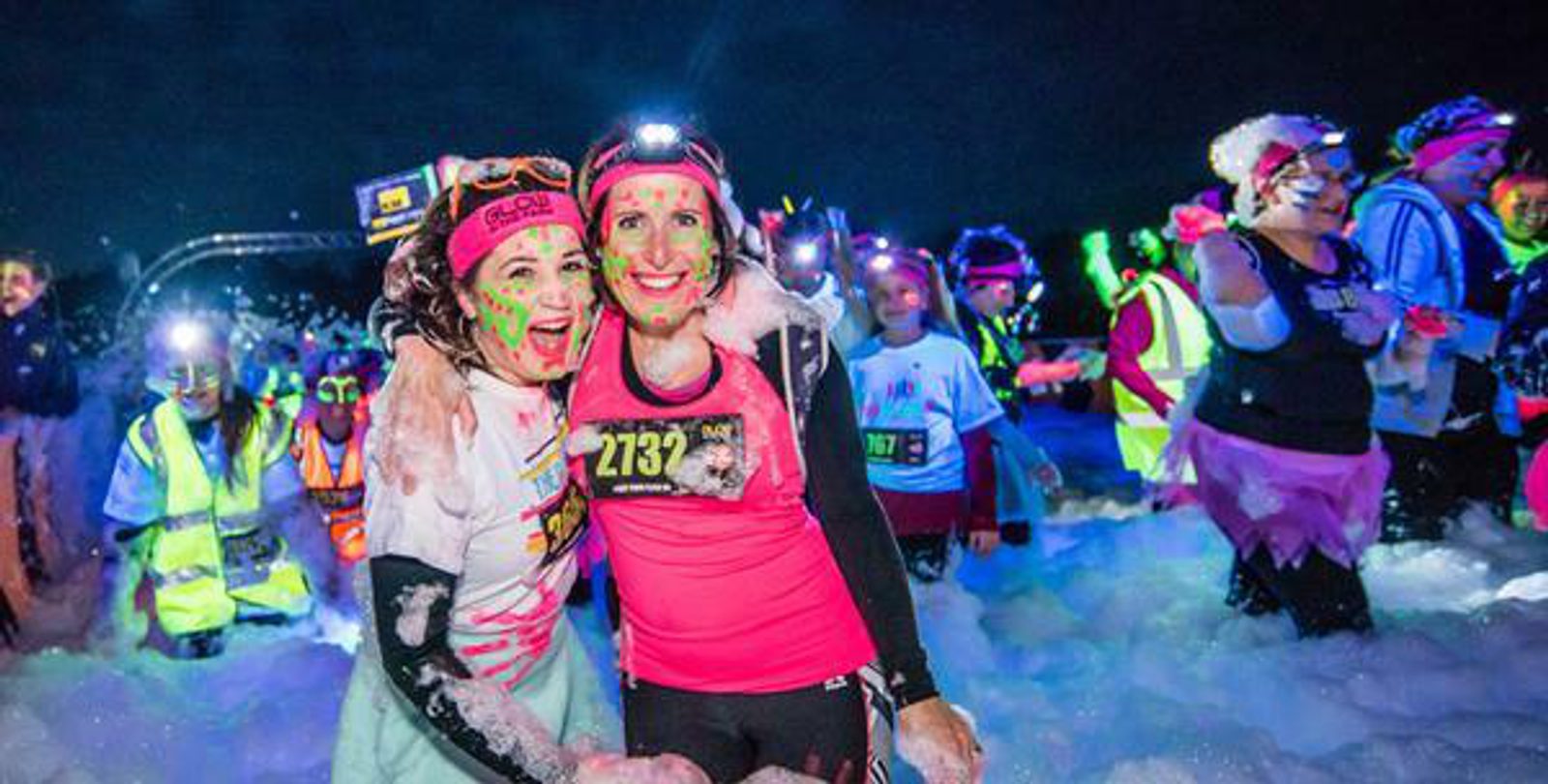 A group of people, many dressed in vibrant and neon clothing, participate in a nighttime foam run. Two women in the foreground, covered in foam and smiling, pose together. They wear headlamps, race bibs, and have glow-in-the-dark paint on their faces and clothes.