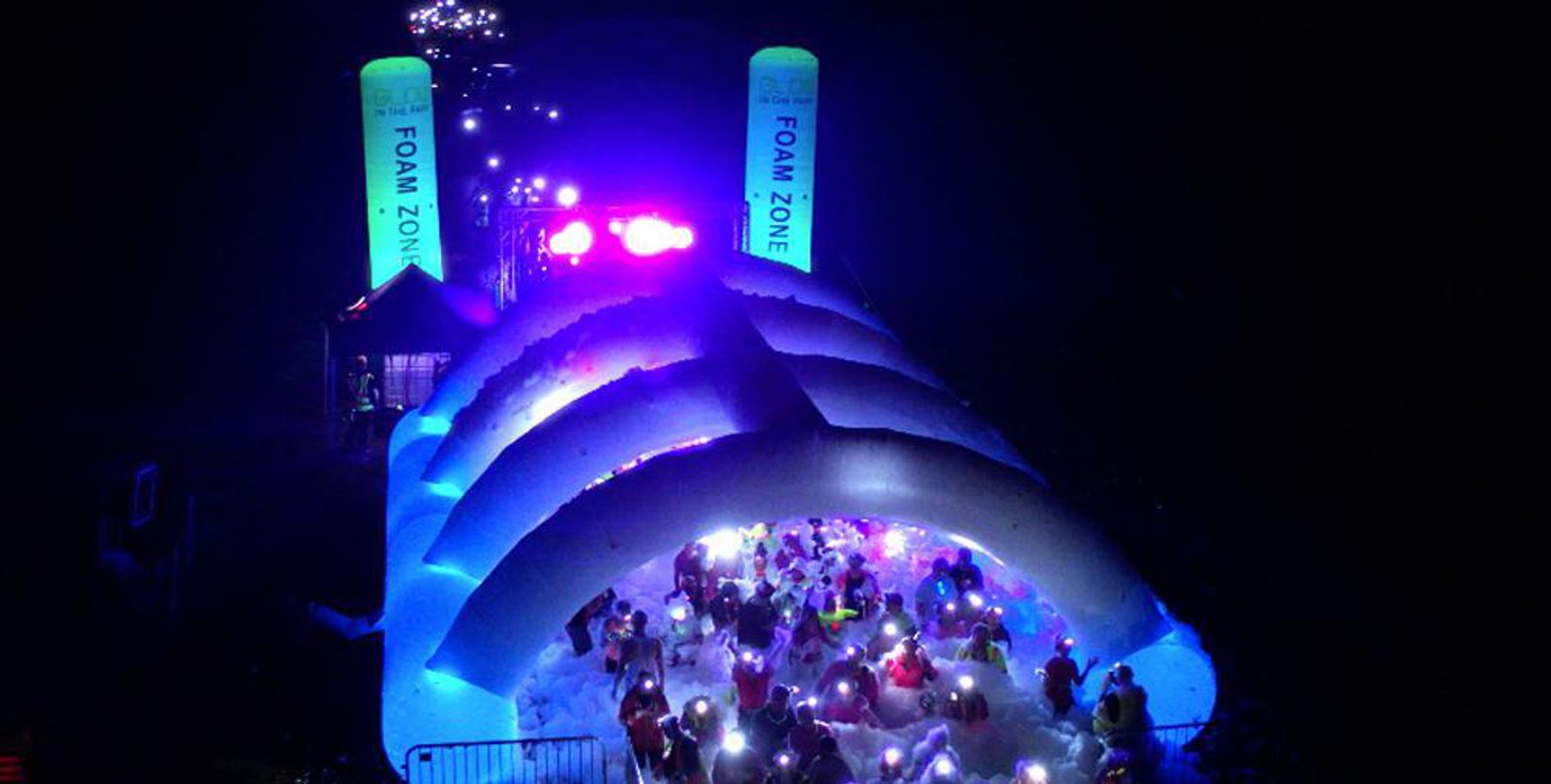 Nighttime image of a "Foam Zone" event with people illuminated by colorful lights. The scene shows a large inflatable structure filled with foam and bubbles, while participants, wearing glow accessories and running gear, navigate through the foamy area under bright lighting.