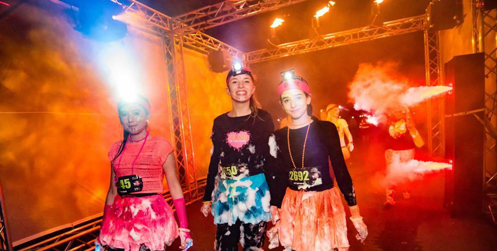 Three participants wearing running numbers and neon clothes are smiling during a nighttime event. They have headlamps and neon accessories, with illuminated backgrounds and smoke effects. The scene appears vibrant and festive.