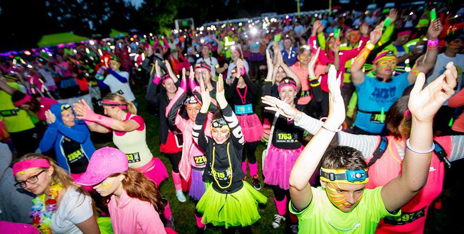 A large group of people are gathered outdoors at dusk, participating in a vibrant event. Many are dressed in neon-colored clothing and accessories, including tutus and headbands. Some have face paint and glow sticks, with their arms raised in celebration.