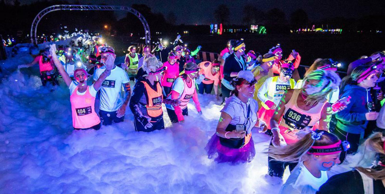 A large group of runners wearing neon clothing and headlamps participate in a nighttime foam run. The participants are surrounded by thick foam, colorful lights, and appear to be enjoying the energetic and vibrant atmosphere.
