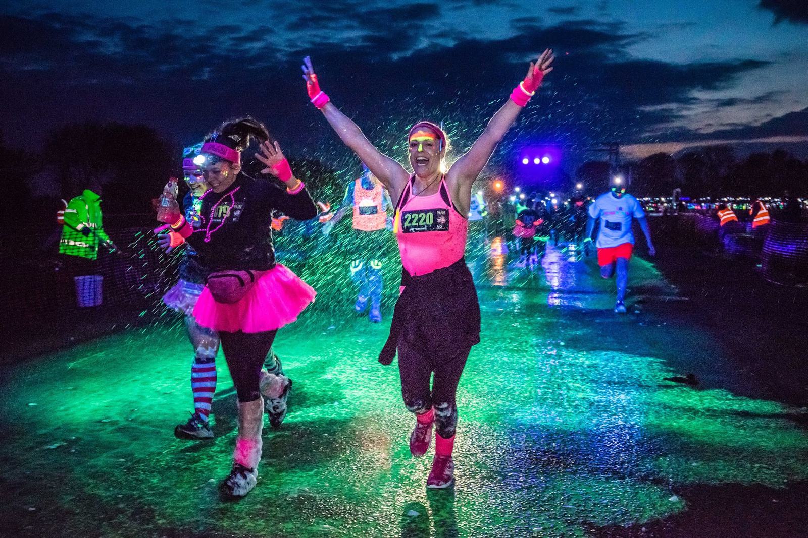 Participants in colorful outfits, including neon lights and tutus, run and walk during a night-time event. One woman with race bib "2220" raises her arms triumphantly, surrounded by runners on a wet, illuminated path with a dark sky in the background.