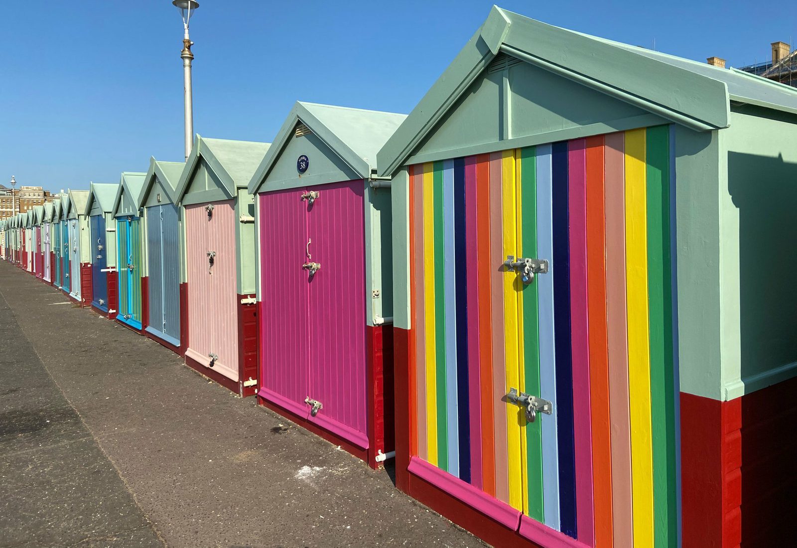 A row of beach huts in various colors stands under a clear blue sky. The huts feature pastel shades such as blue, pink, and mint green. One hut is particularly vibrant, decorated with vertical rainbow stripes. A lamppost is visible in the background.
