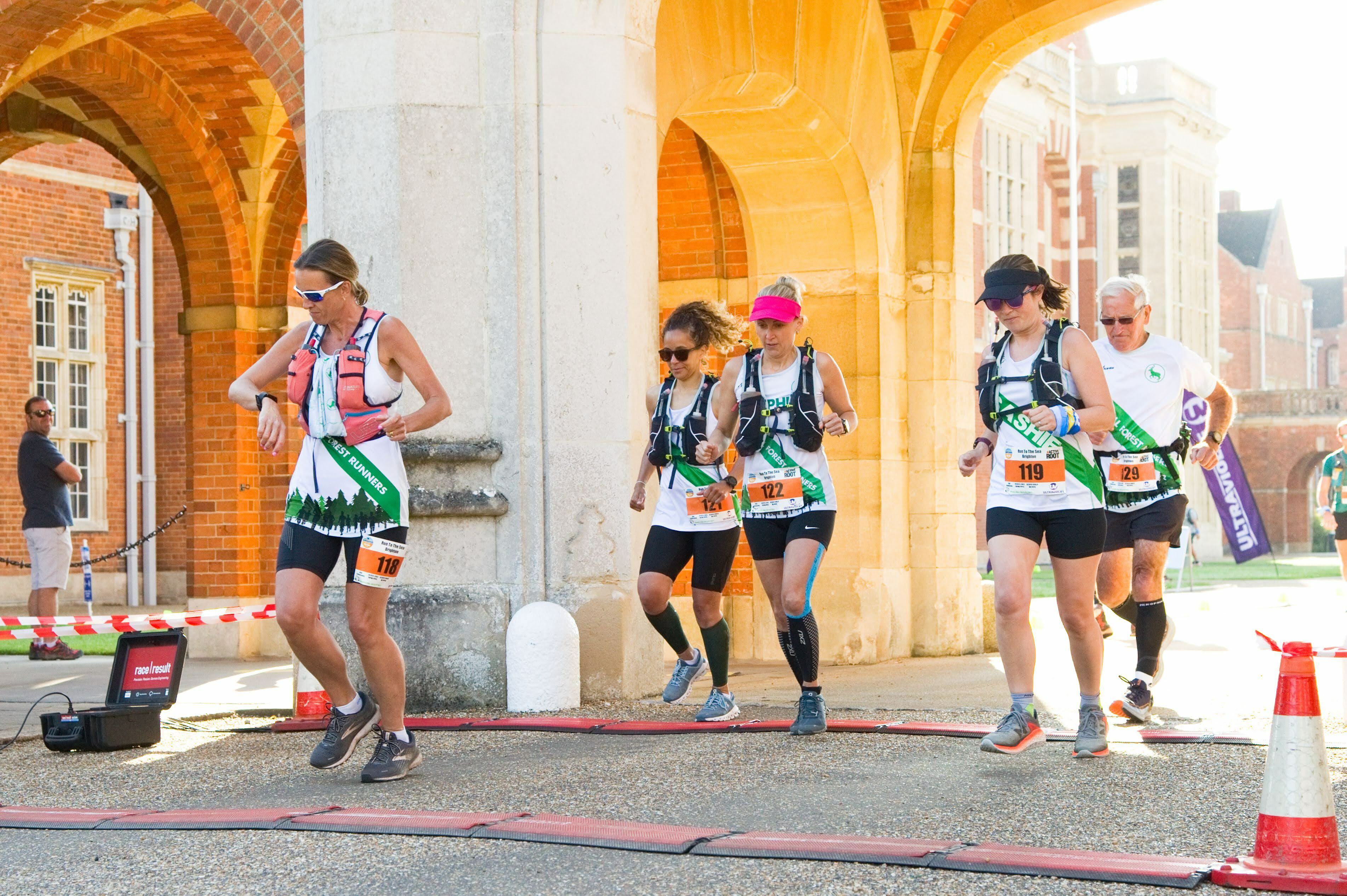 A group of runners crosses the starting line during a race. The sun shines brightly on the brick buildings in the background. Each runner wears a bib and gear, with one person adjusting their clothing. Red and white barriers line the path.