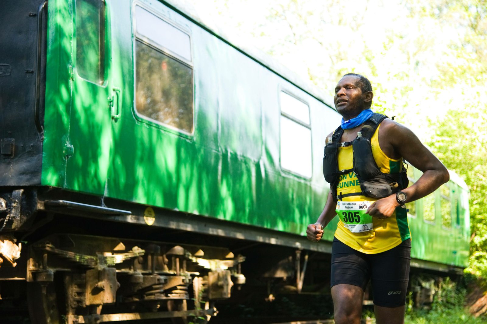 A man wearing a yellow and green tank top with the number 305 runs past an old, green train carriage on a sunny day. He is equipped with a hydration backpack and appears focused on the trail. The background is lush with greenery.