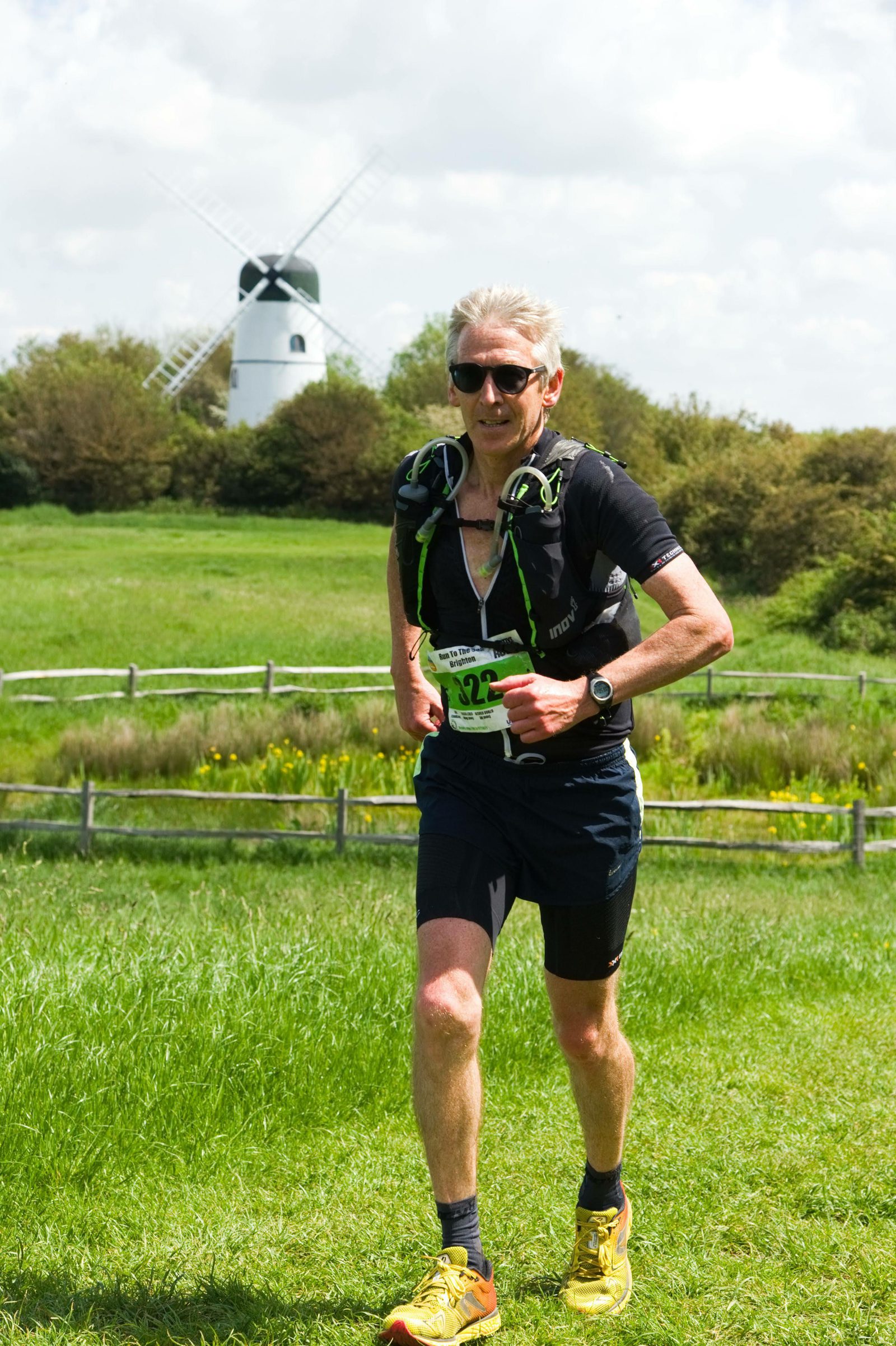 A runner with short blonde hair, wearing sunglasses, a dark shirt, shorts, yellow shoes, and a hydration pack jogs in a lush green field. A white windmill and trees are in the background under a partly cloudy sky. The runner has a race bib pinned to his shirt.