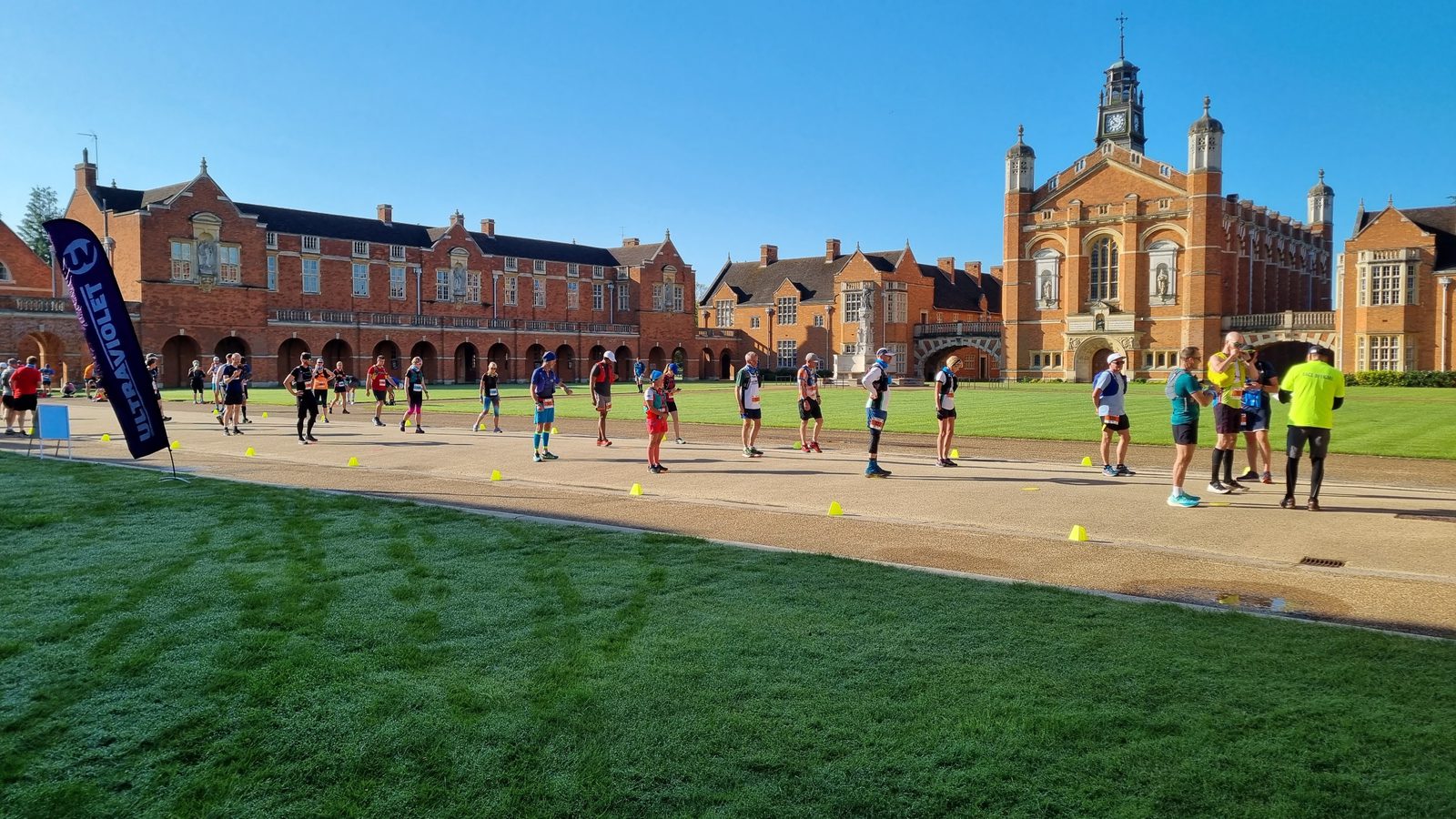 A group of people stand in lines on a paved path marked with yellow cones, set against a backdrop of large, historic red-brick buildings. Some participants wear running attire, and a banner is visible on the left. The sky is clear and blue, indicating sunny weather.