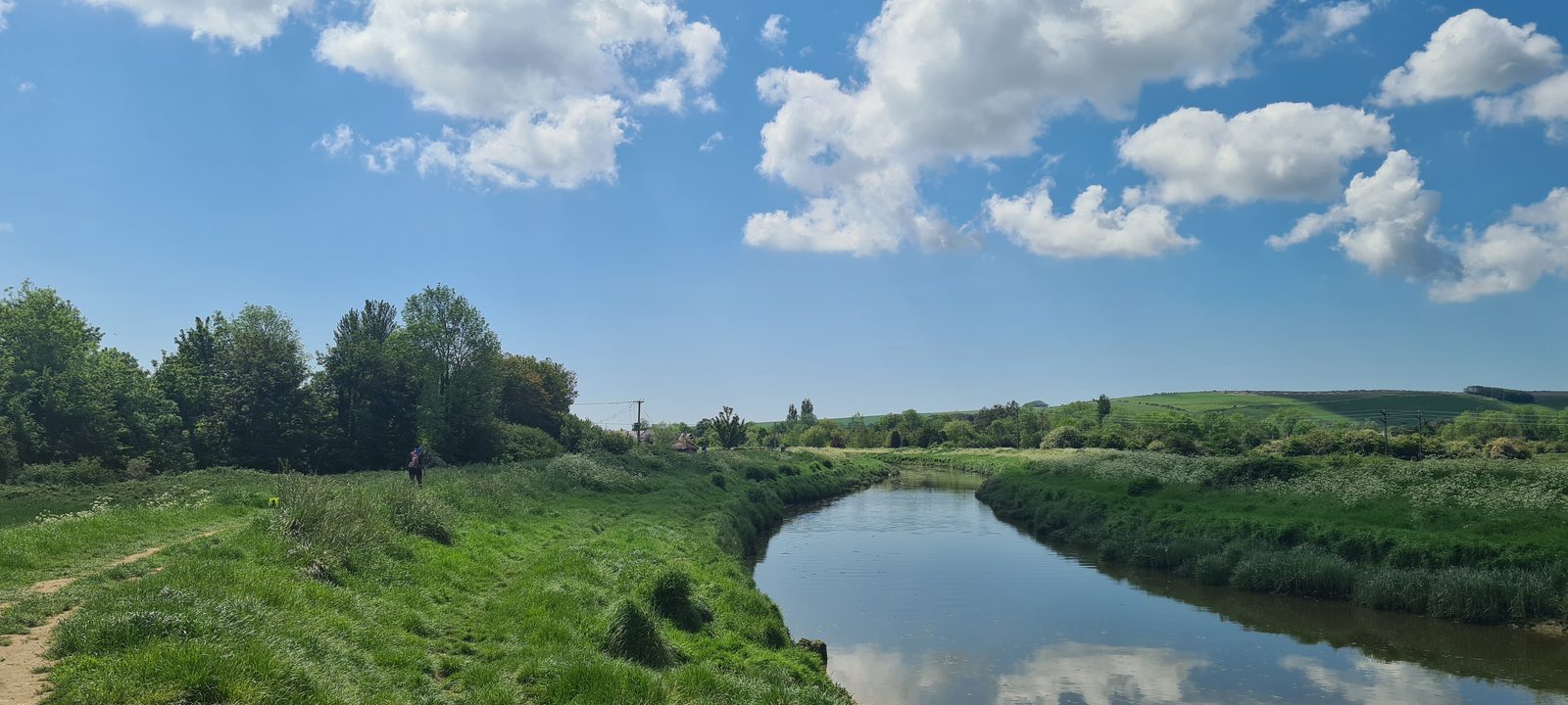 A peaceful riverside scene with a clear, winding river reflecting the blue sky and fluffy white clouds. Lush green grass and trees line the banks, with a dirt path on the left. Rolling hills are visible in the distance under a bright, sunny sky.