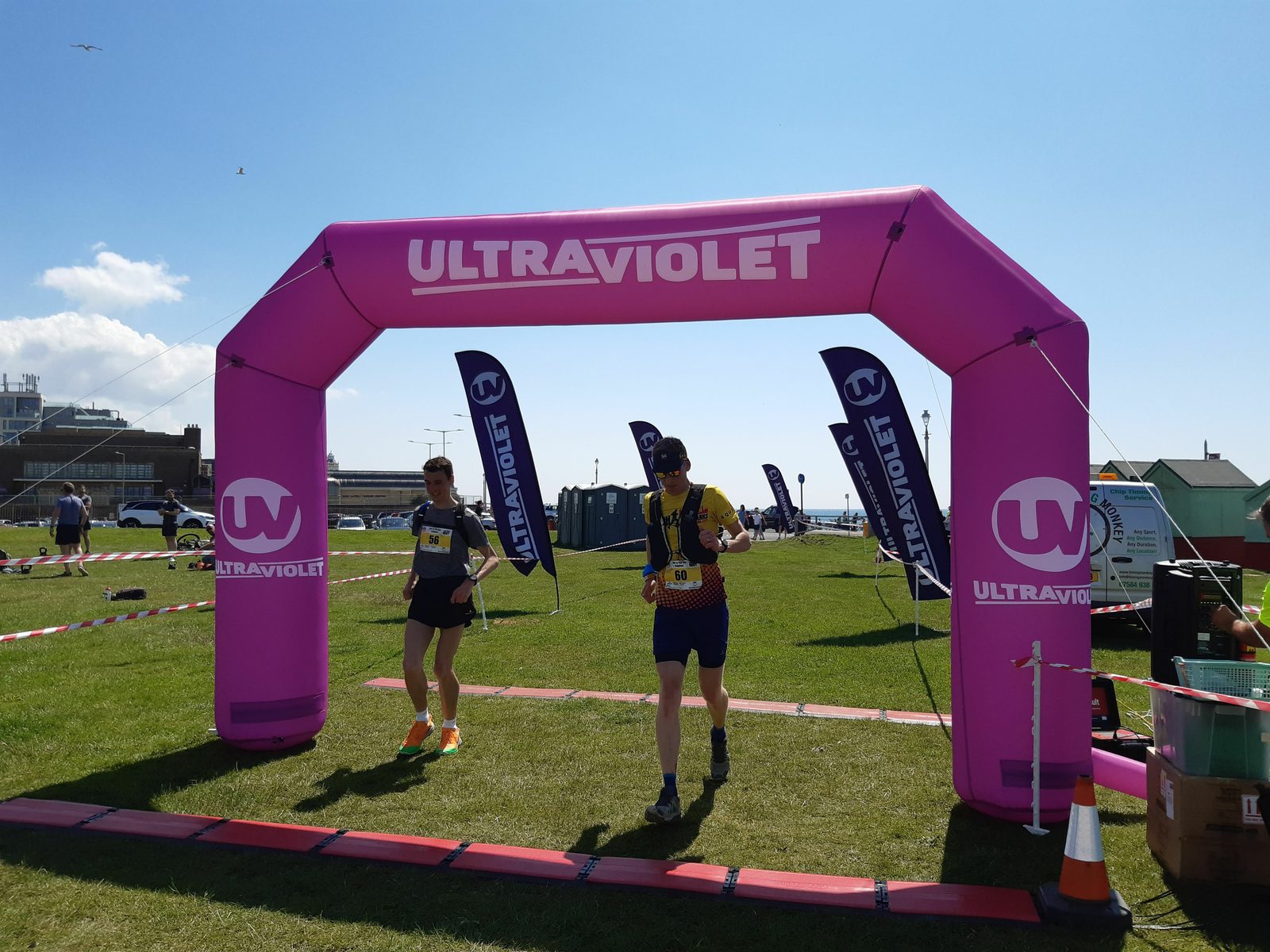 Two runners are crossing the finish line under a pink inflatable arch marked "ULTRAVIOLET" at an outdoor event. The event is set on a grassy field with sky and a few scattered clouds above. Flags with "UV" logos and various event equipment are visible in the background.