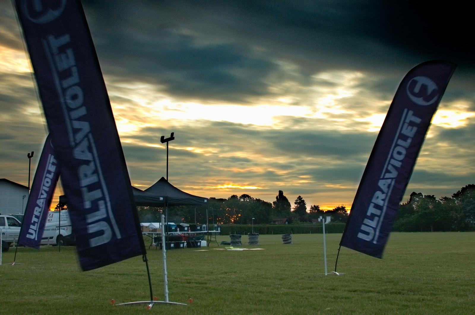 A field at sunset with dark clouds above. Three tall, narrow banners reading "ULTRAVIOLET" are placed on the field. A black canopy tent and equipment are visible in the background, along with trees and a few buildings near the horizon.