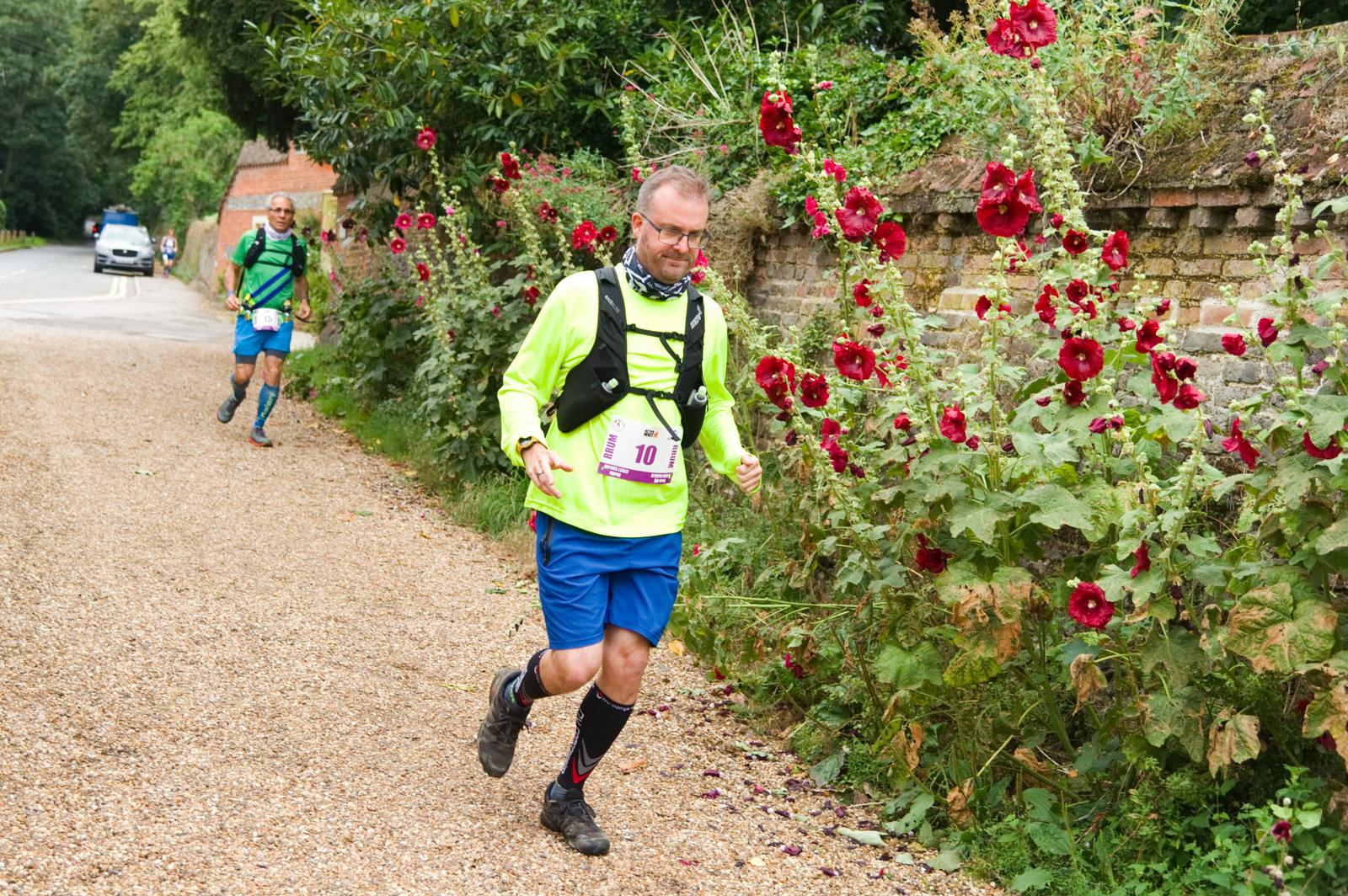 A man in a bright yellow shirt and blue shorts runs along a gravel path lined with tall red flowers and greenery. He wears a race bib numbered 10 and a hydration pack. Another runner is visible in the background. A brick wall and trees are seen along the path.