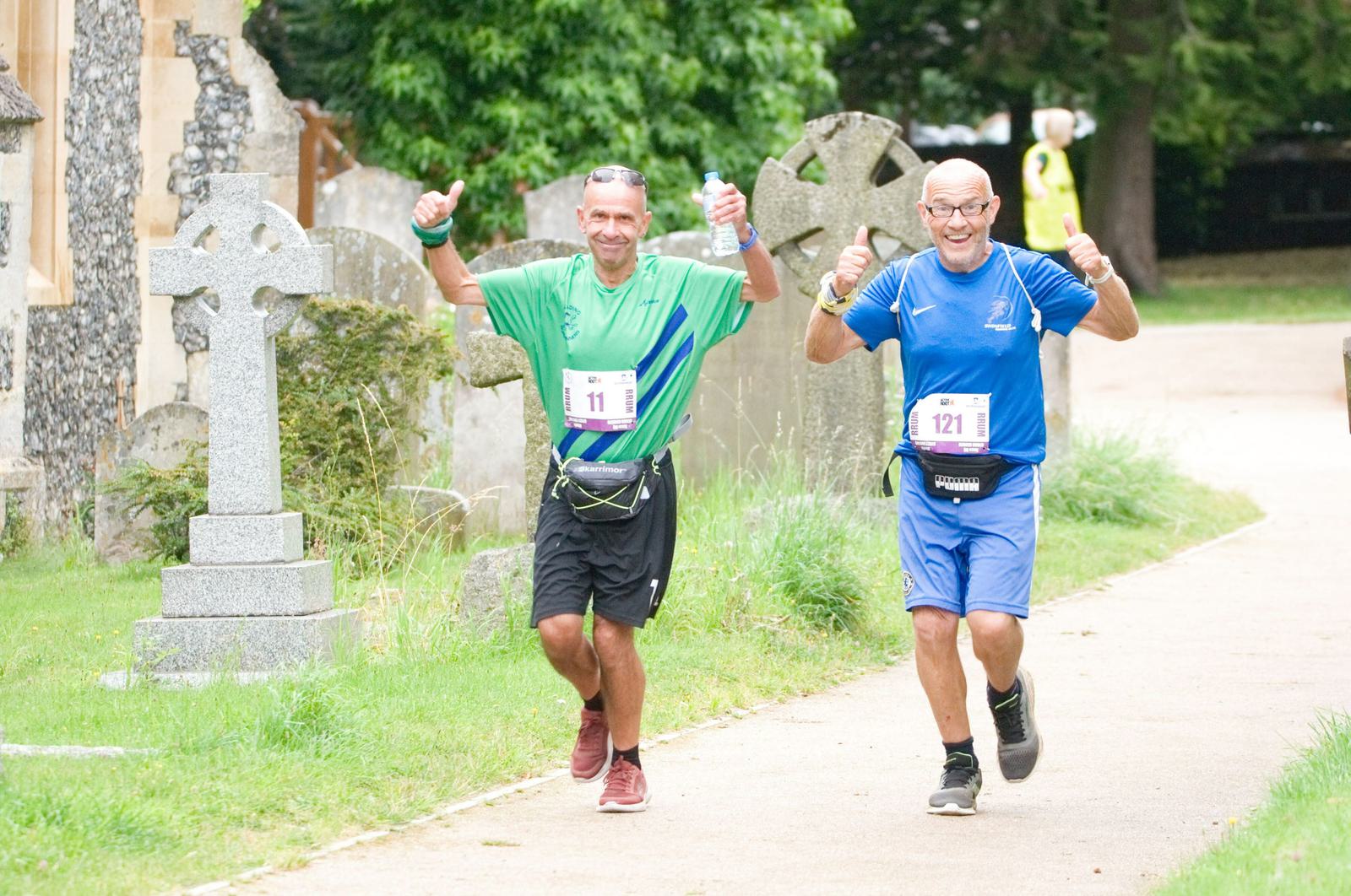 Two men in athletic attire participate in a race, running joyfully on a path through a cemetery. Both are wearing race bibs and sneakers, raising their thumbs up in excitement. The man on the left wears a green shirt, while the right one is in blue.