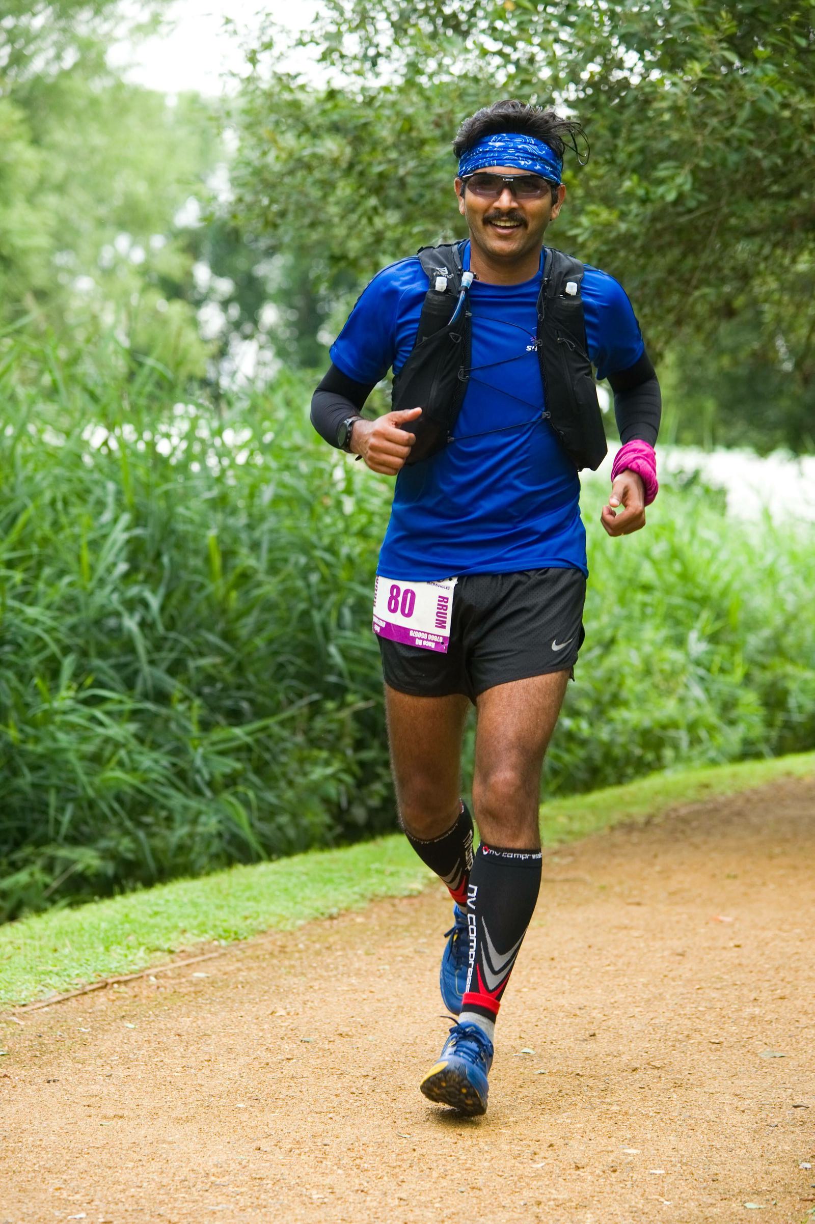A runner in a blue shirt, black shorts, and sports gear, including a headband and arm sleeves, is mid-stride on a dirt path surrounded by lush greenery. A race bib numbered 80 is attached to his shorts. A cheerful, focused expression is visible on his face.