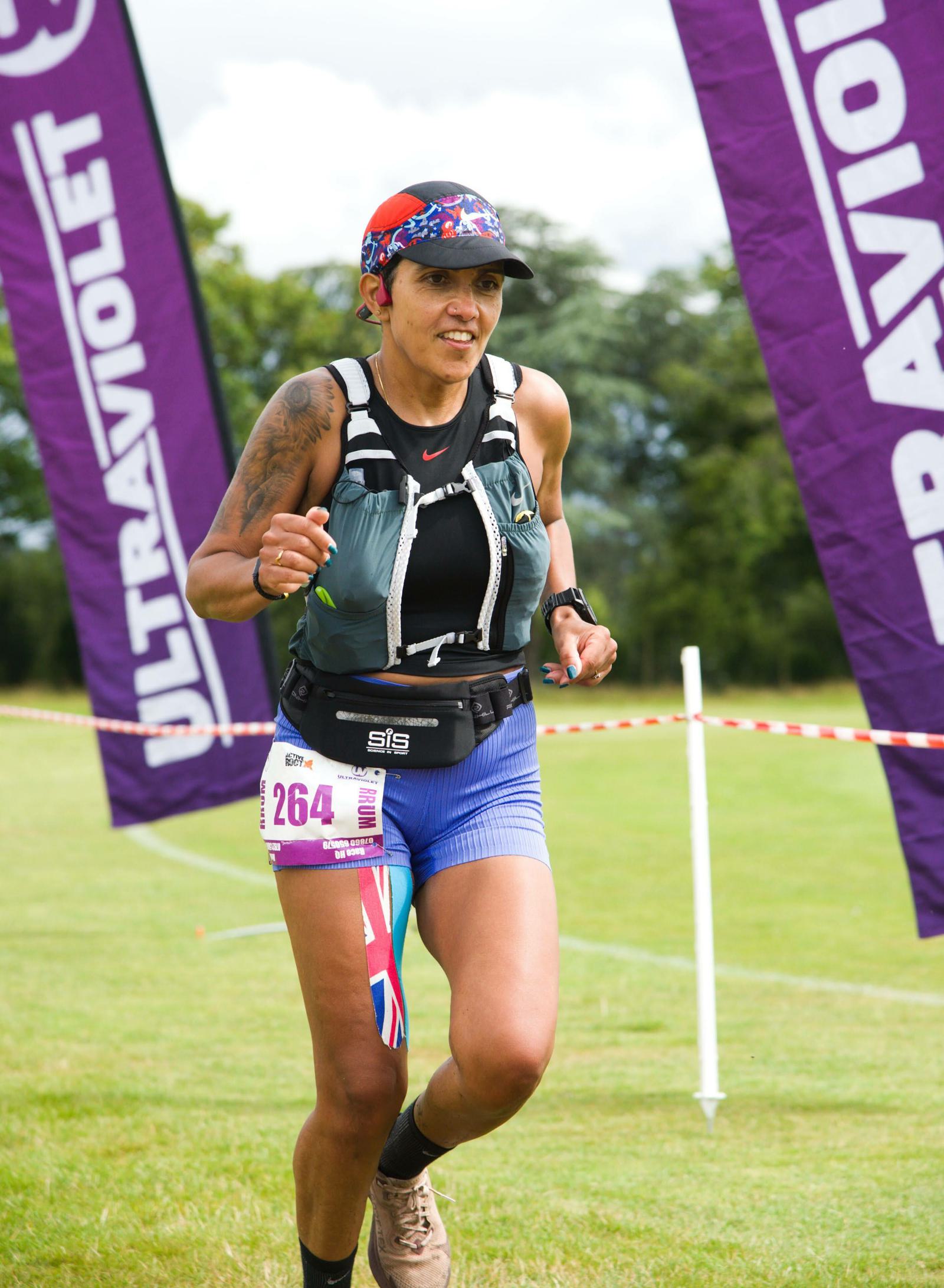 A runner wearing a cap and hydration vest competes in a race, running past purple banners that read "ULTRAVIOLET." The runner is sporting a pink race number 264 on their shorts, while the background features green grass and trees.