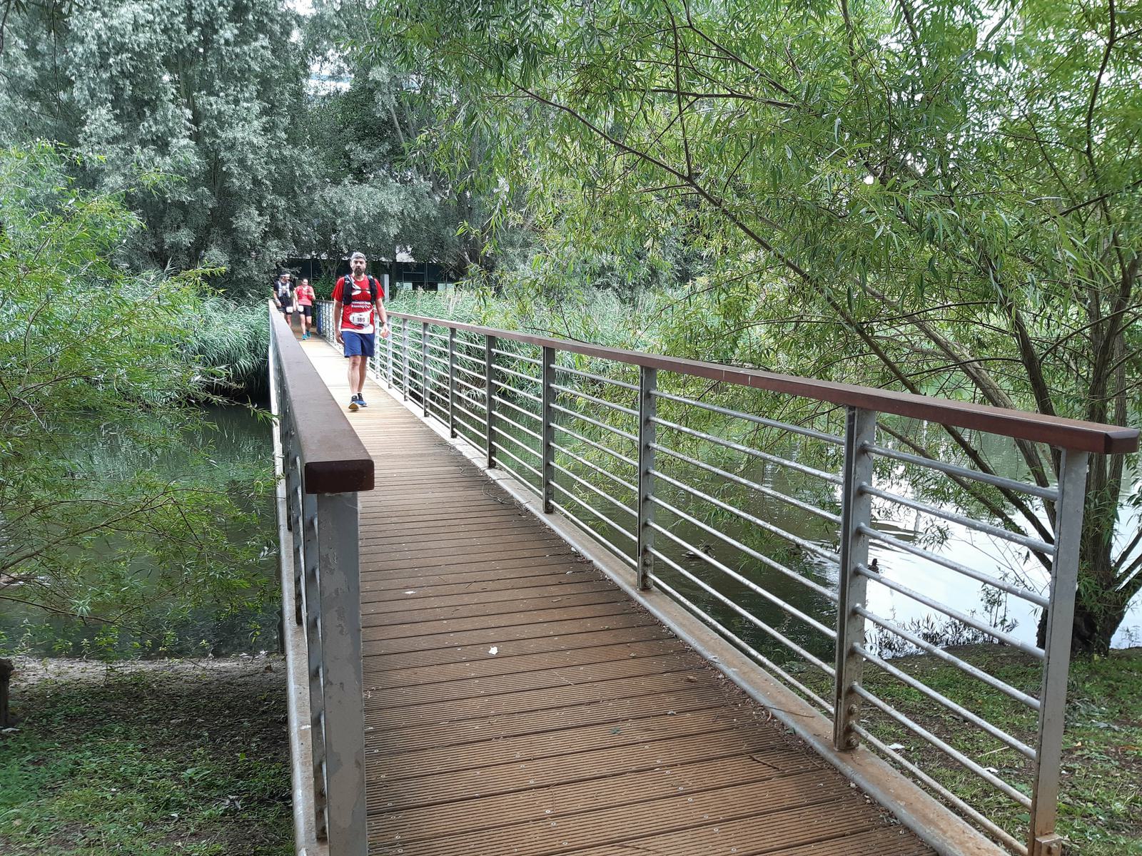 A wooden footbridge with metal railings spans a lush, green riverbank. A few people walk along it, including a person in a red shirt and white shorts heading towards the camera. Trees with dense foliage surround the bridge, creating a serene nature scene.
