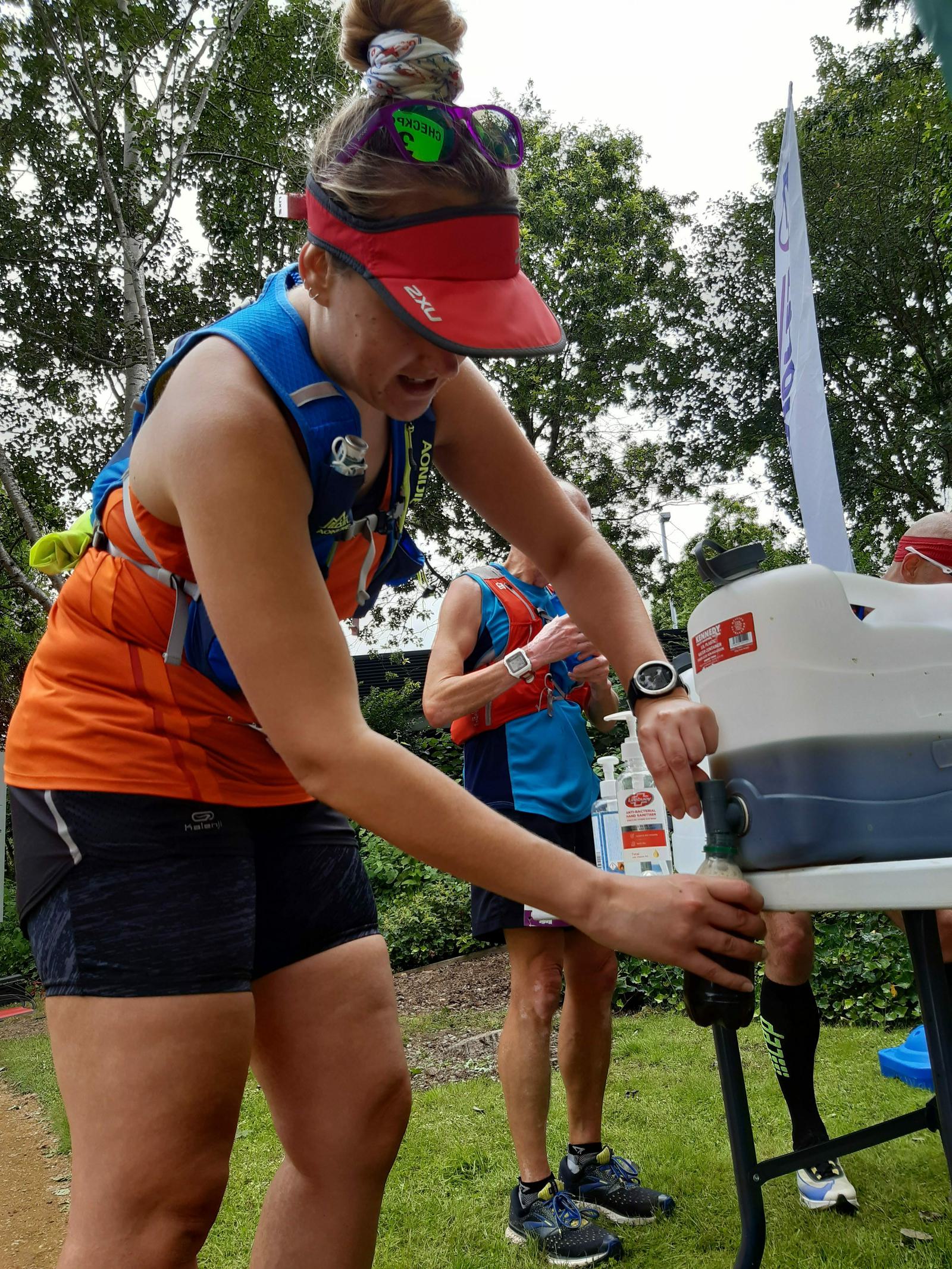 A runner wearing an orange vest, sunglasses, and a red visor fills a water bottle from a dispenser at a hydration station during a race. Another runner in blue stands behind her drinking from a bottle. Trees and greenery are visible in the background.