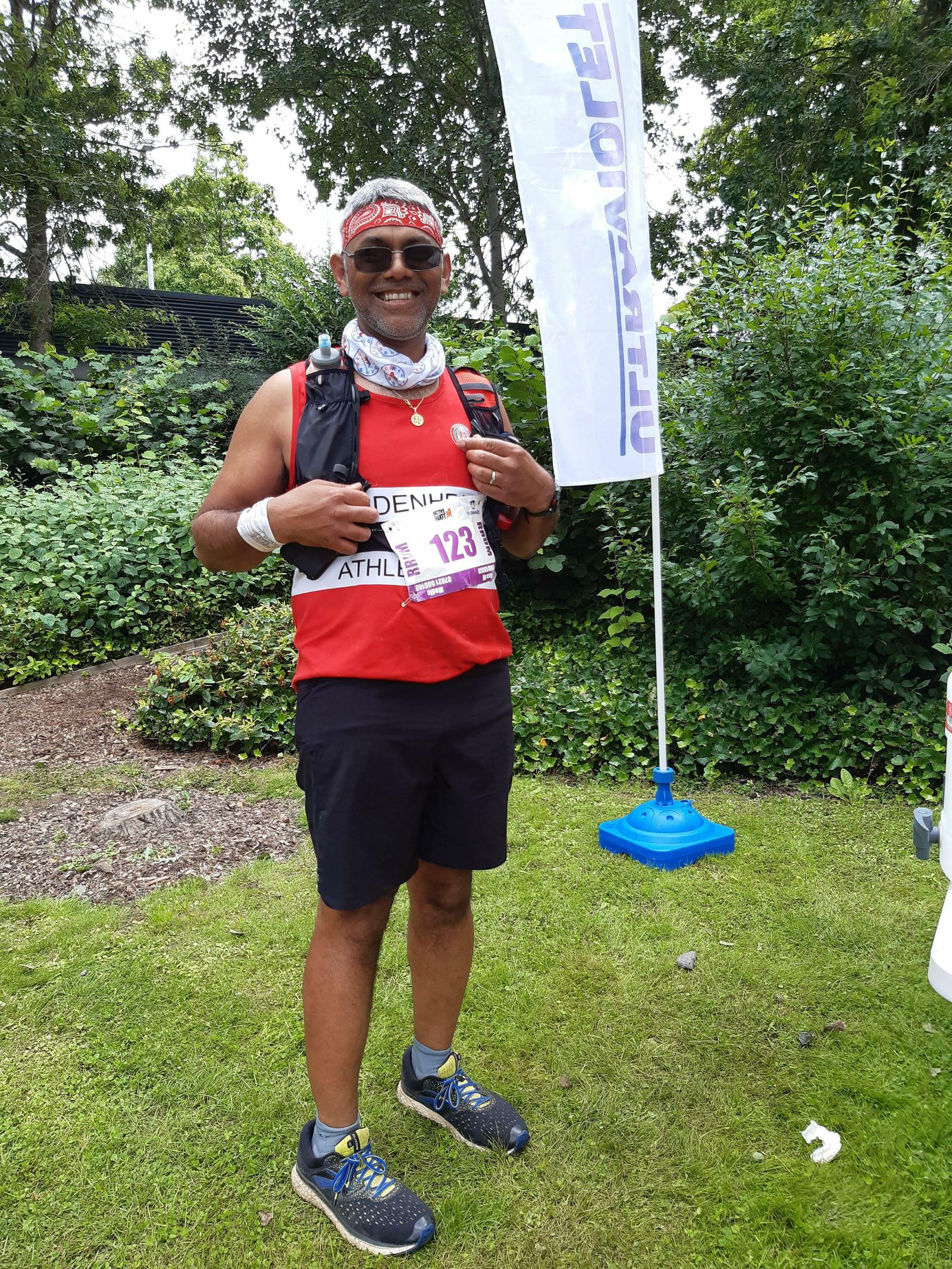 A man stands on grass in a park after completing a race. He wears a red sleeveless shirt, black shorts, and a race bib numbered 123. He has a bandana on his head, sunglasses, and a neck gaiter. He smiles and makes a thumbs-up gesture. Trees and a sign are in the background.