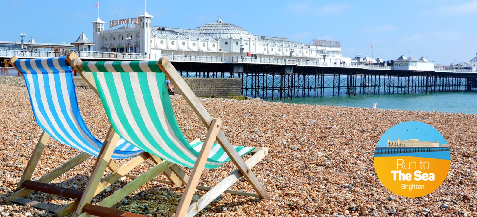Two striped deck chairs sit on a pebbled beach with a view of Brighton Pier in the background. The bright blue sky and calm sea add to the scenic coastal setting. An inset circle with a graphic of the pier reads "Run to The Sea Brighton.
