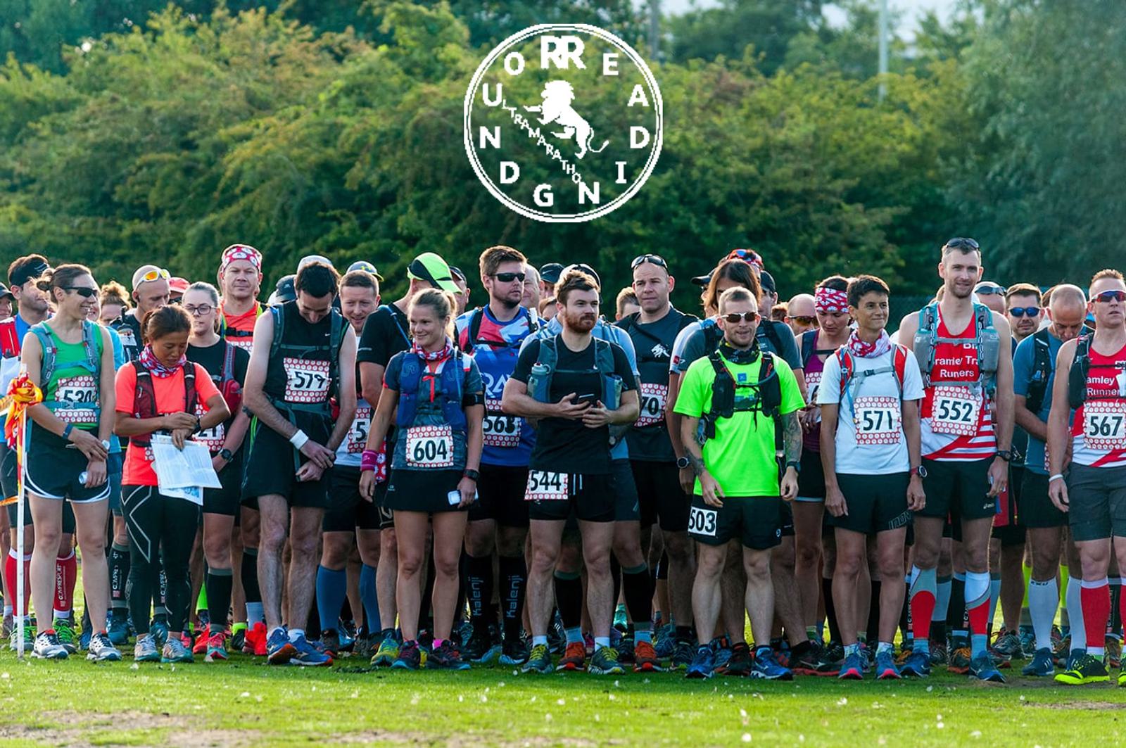 A diverse group of runners stands at the starting line of a race, ready to begin. They wear race bibs and running gear, and some carry backpacks. The surroundings are green and sunlit, with a logo containing various alphabet letters at the top of the photo.