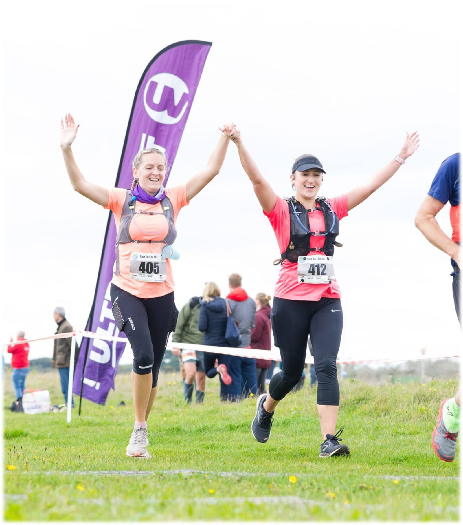 Two smiling women in running gear cross the finish line of a race, holding hands and raising their arms in celebration. Both wear numbered bibs (405 and 412). A purple flag and several onlookers are visible in the background.
