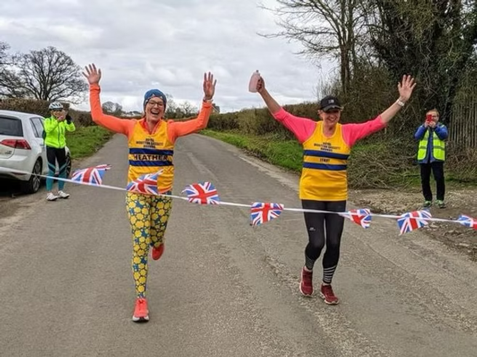 Two smiling runners in colorful outfits cross a finish line adorned with Union Jack flags. They raise their arms in victory on a rural road, while a few onlookers and another runner cheer them on.