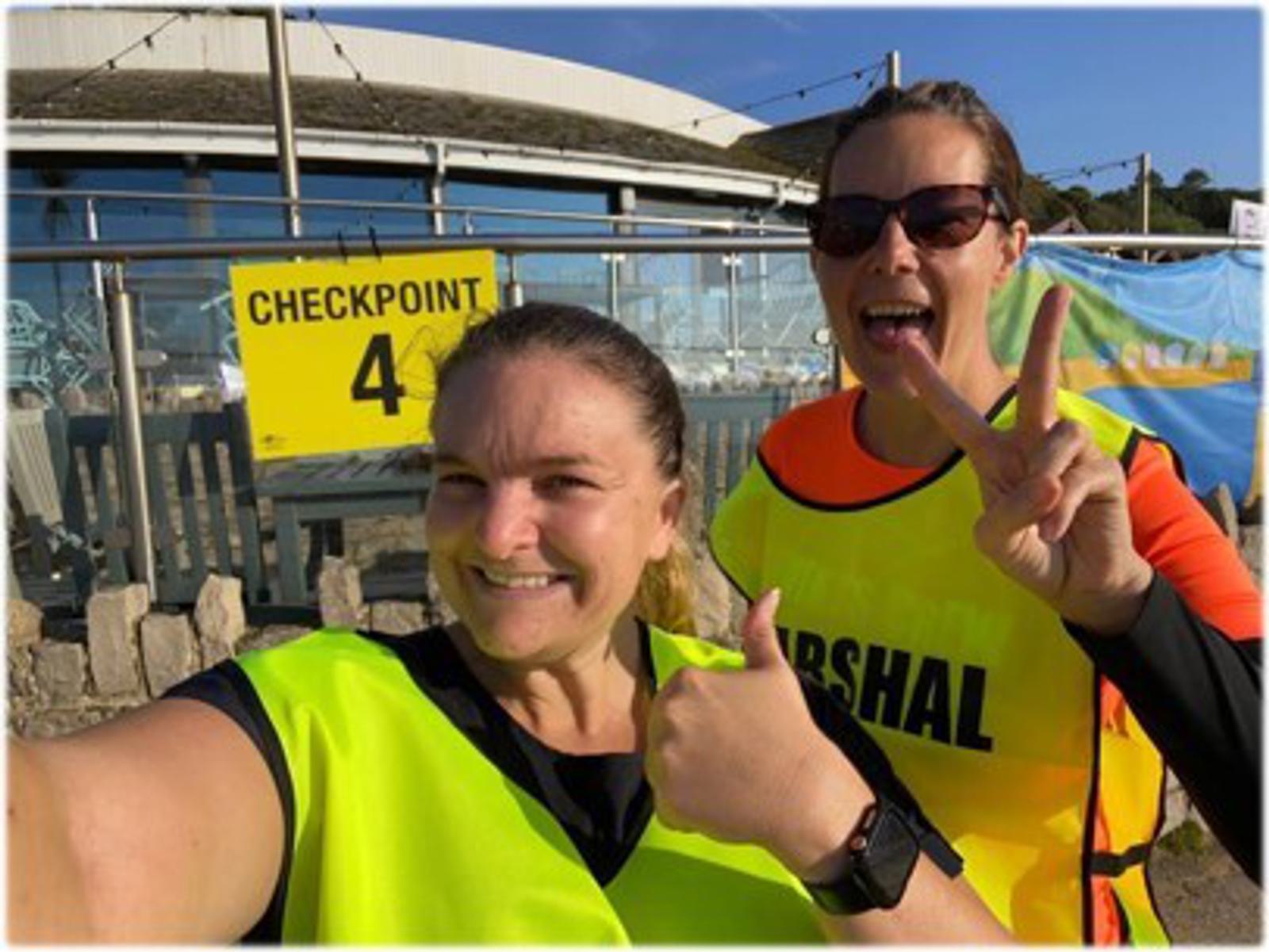 Two people in fluorescent vests smile at the camera near a "Checkpoint 4" sign. One gives a thumbs up, the other holds up a peace sign. They appear to be outdoors on a sunny day.