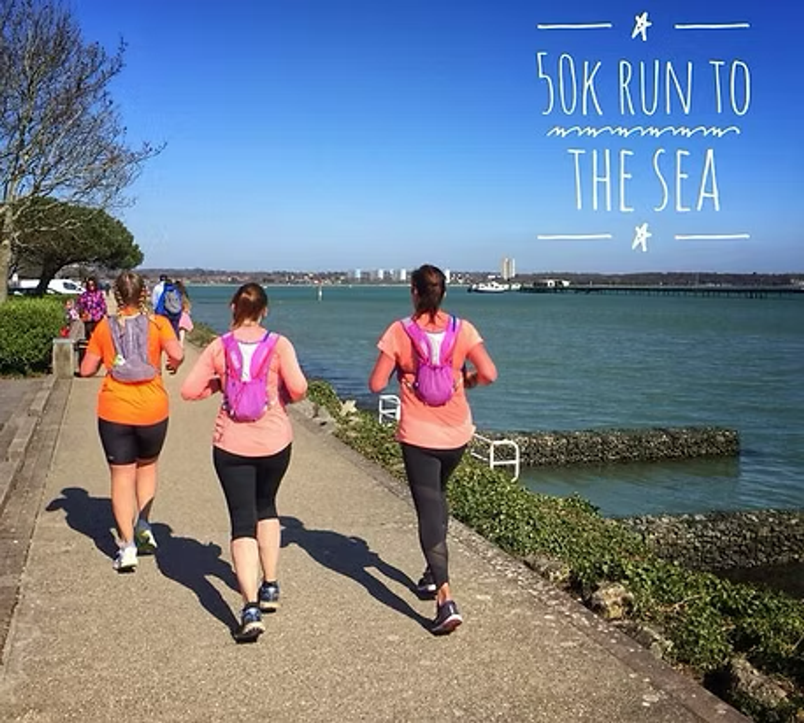 Three people jogging along a seaside path under a clear blue sky. They each wear running gear and backpacks. The water is calm, and the skyline is visible in the distance. Text reads "50K Run to the Sea.