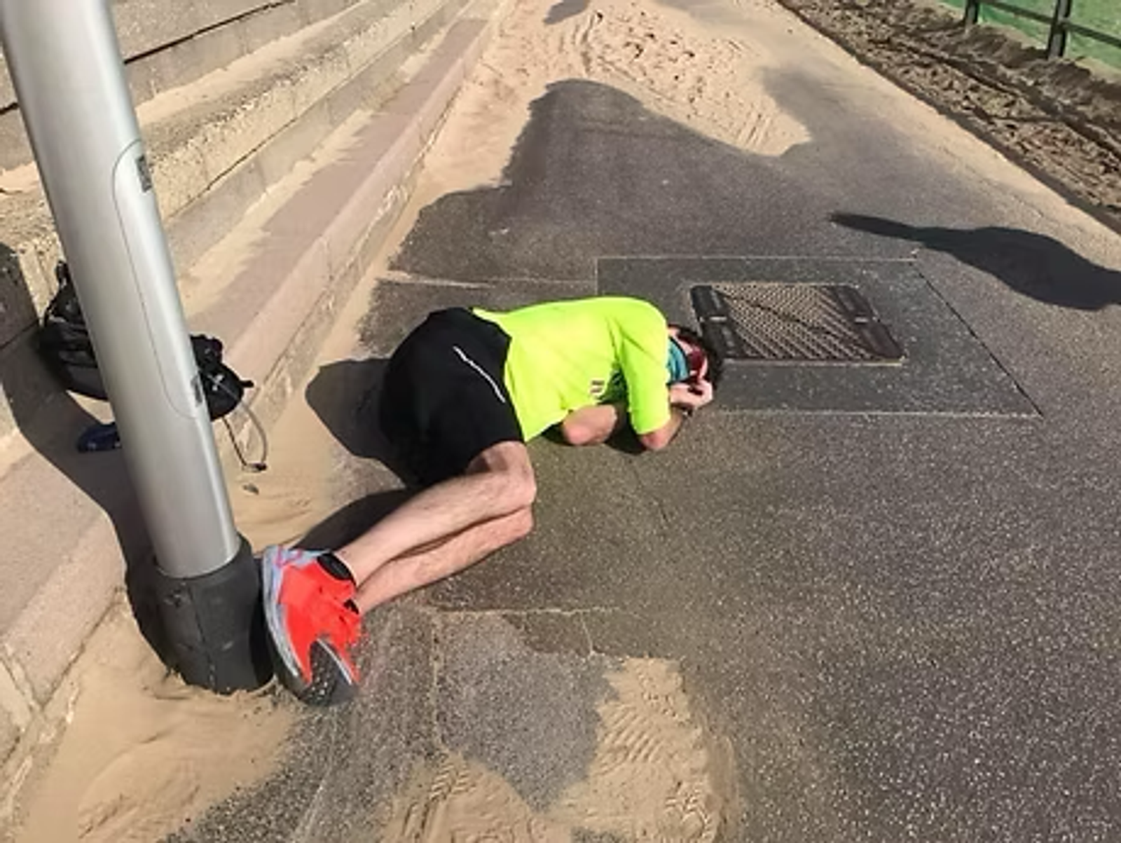 A person in a neon green shirt and red shoes is lying on the ground near a lamp post, holding their head. They are on a path with sand scattered around.