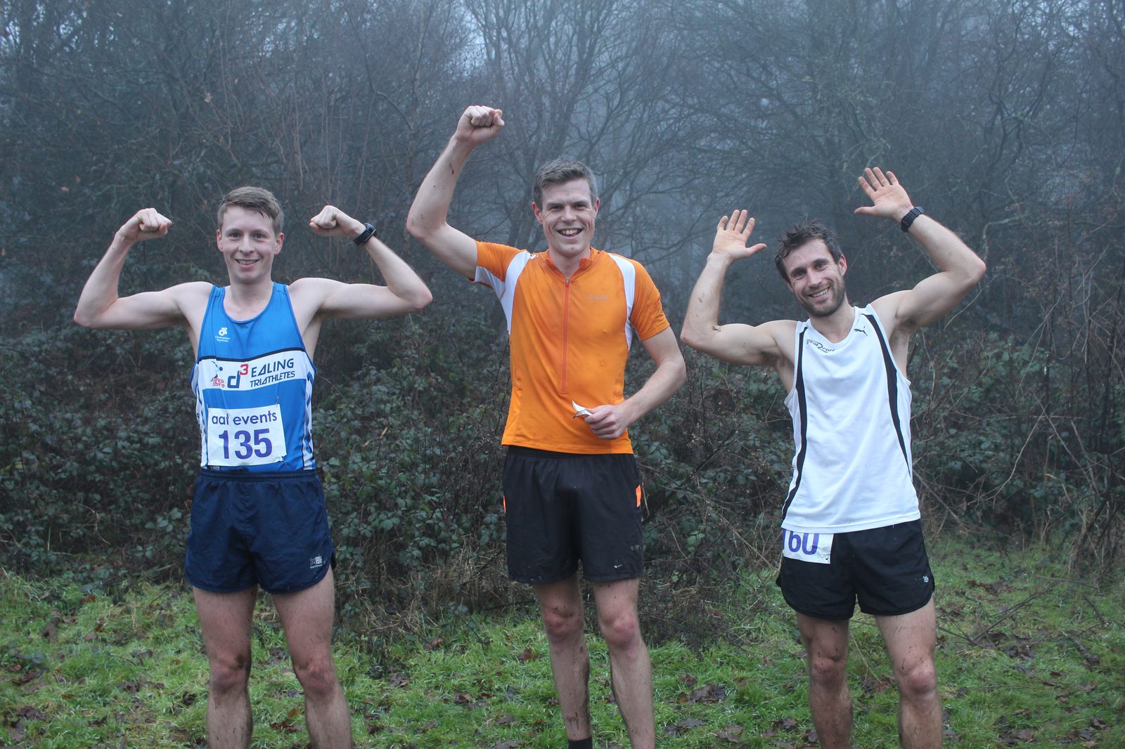 Three runners pose, flexing their biceps and smiling after completing a race. The runner on the left is in blue, the center runner in orange, and the runner on the right in white. They stand in a grassy area with leafless trees and a foggy backdrop.