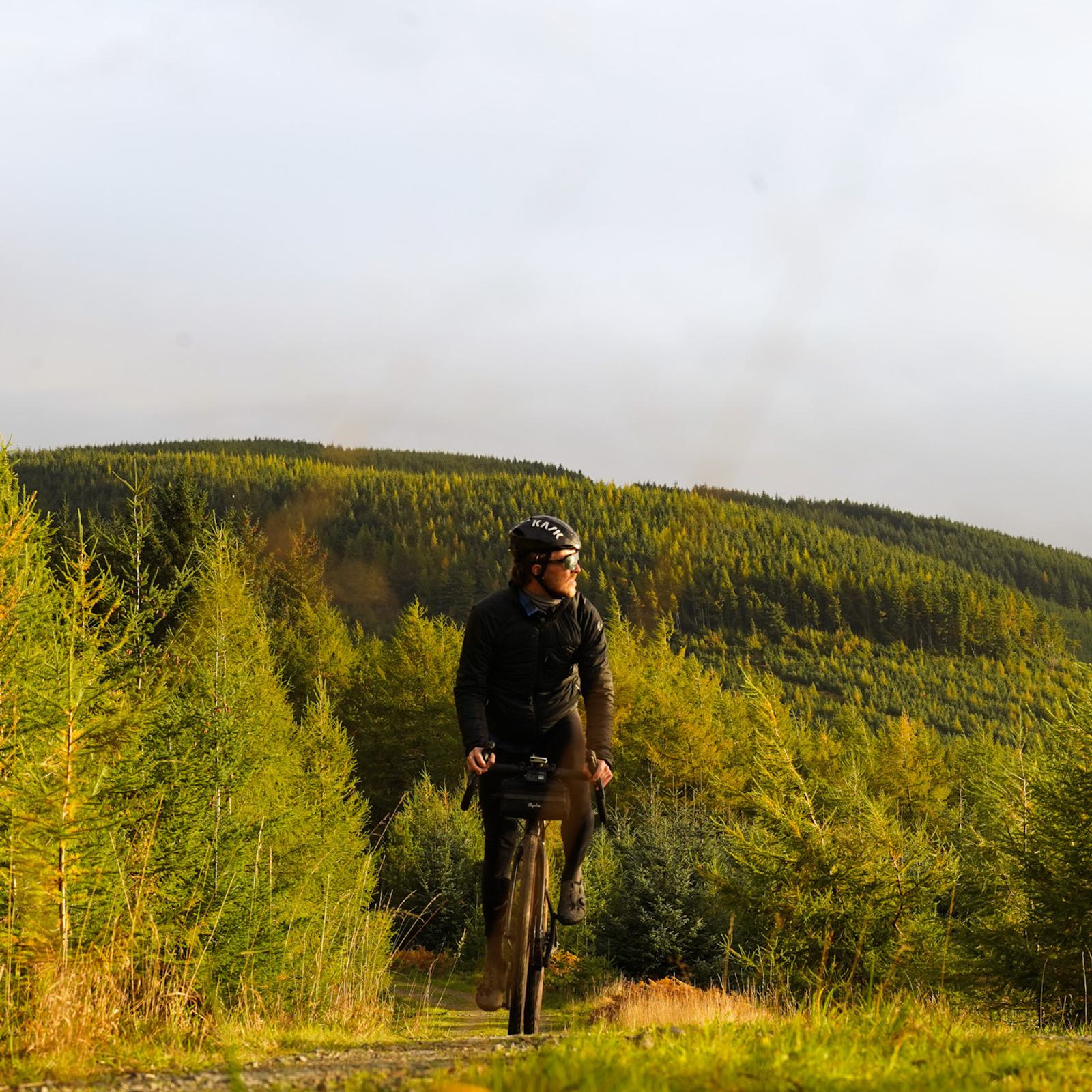 A person wearing a black cycling outfit and helmet rides a bike on a path surrounded by lush, green trees and foliage. The background reveals rolling hills covered in dense forest under a cloudy sky.