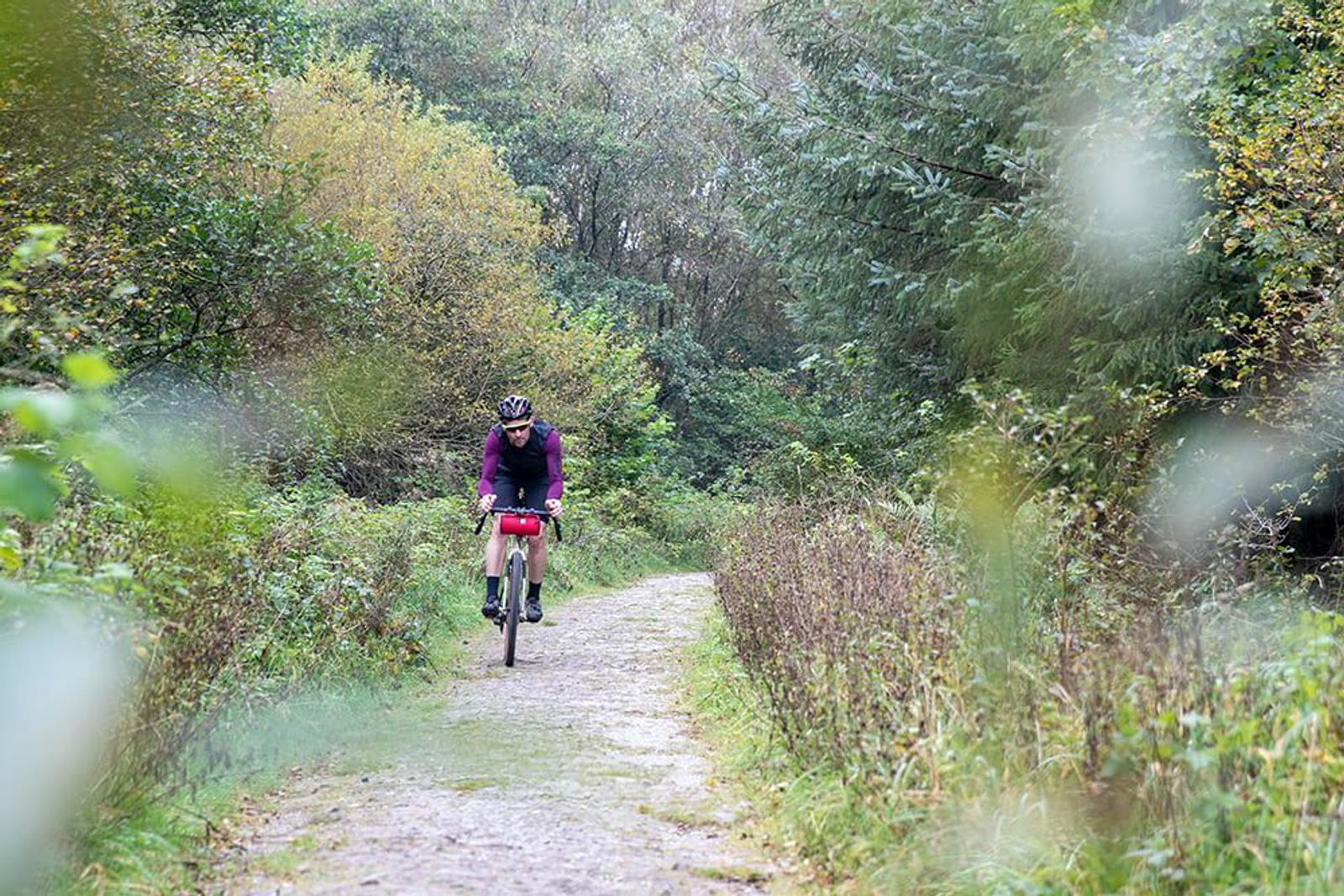 A cyclist wearing a helmet and casual biking gear rides along a gravel path through a lush, green forest. The surrounding trees and foliage create a peaceful, natural atmosphere. Light filters through the leaves, giving the scene a serene and refreshing feel.