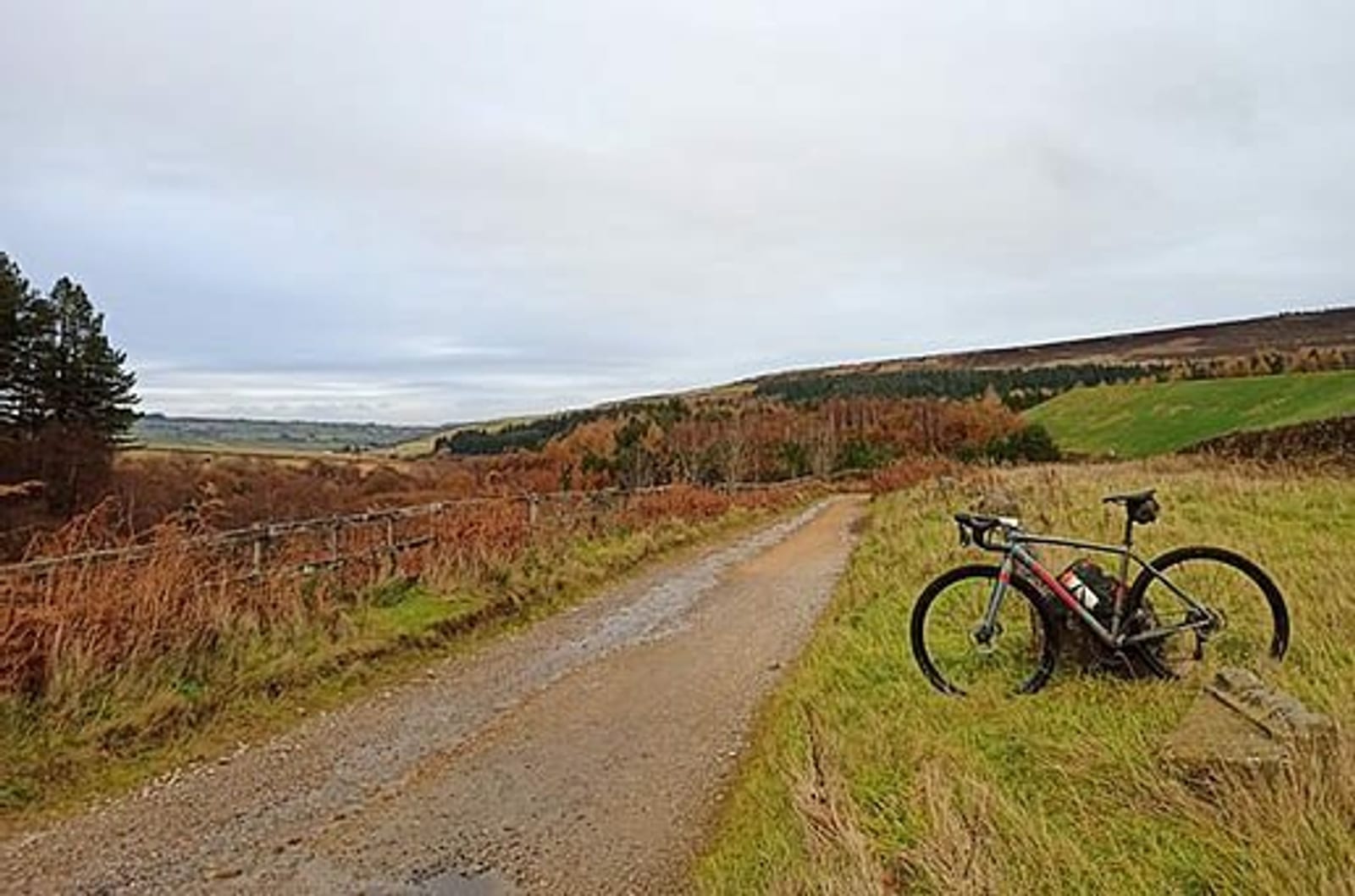 A gravel path winds through a scenic, hilly landscape covered with patches of green grass, trees, and brown foliage. A bicycle rests on its side near the edge of the path. The sky is overcast, adding a serene atmosphere to the countryside setting.