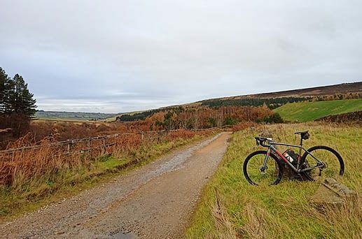 A gravel path winds through a scenic, hilly landscape covered with patches of green grass, trees, and brown foliage. A bicycle rests on its side near the edge of the path. The sky is overcast, adding a serene atmosphere to the countryside setting.