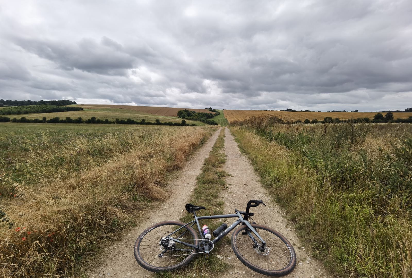 A gravel bike lies on a dirt path flanked by grassy fields and rolling hills. The sky is overcast with dense clouds, creating a moody atmosphere. The dirt path leads to the horizon, offering a serene and scenic view of the countryside.