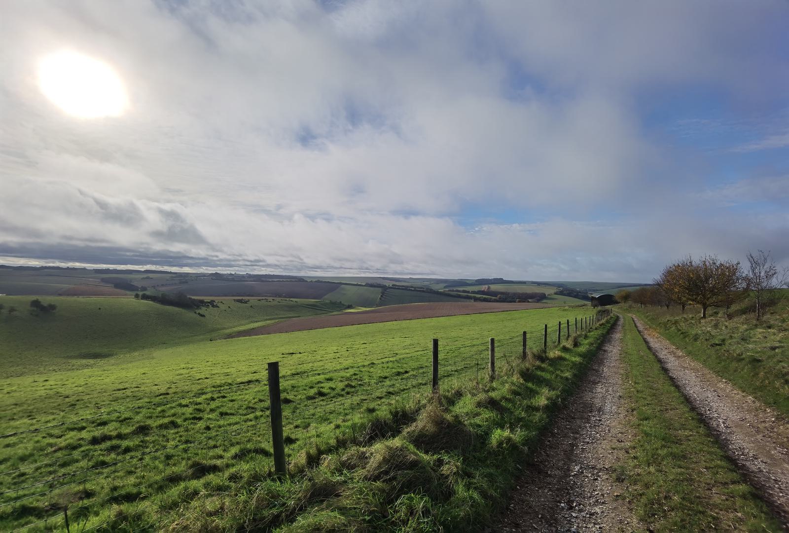 A scenic countryside view showing a gravel path alongside a fence leading into rolling green hills and fields under a cloudy sky with the sun breaking through. Trees and shrubs are scattered across the landscape with distant hills stretching into the horizon.
