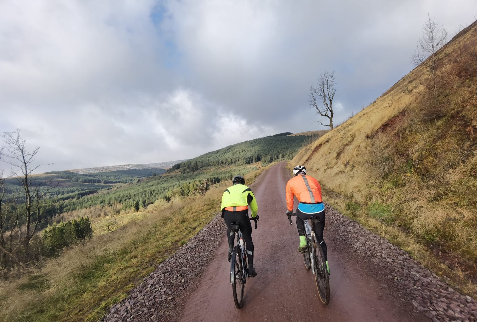 Two cyclists are riding on a gravel path through a scenic forested landscape. They wear bright jackets, one in neon yellow and the other in a mix of orange and blue. The path winds through rolling hills under a partly cloudy sky. Sparse trees flank the path.