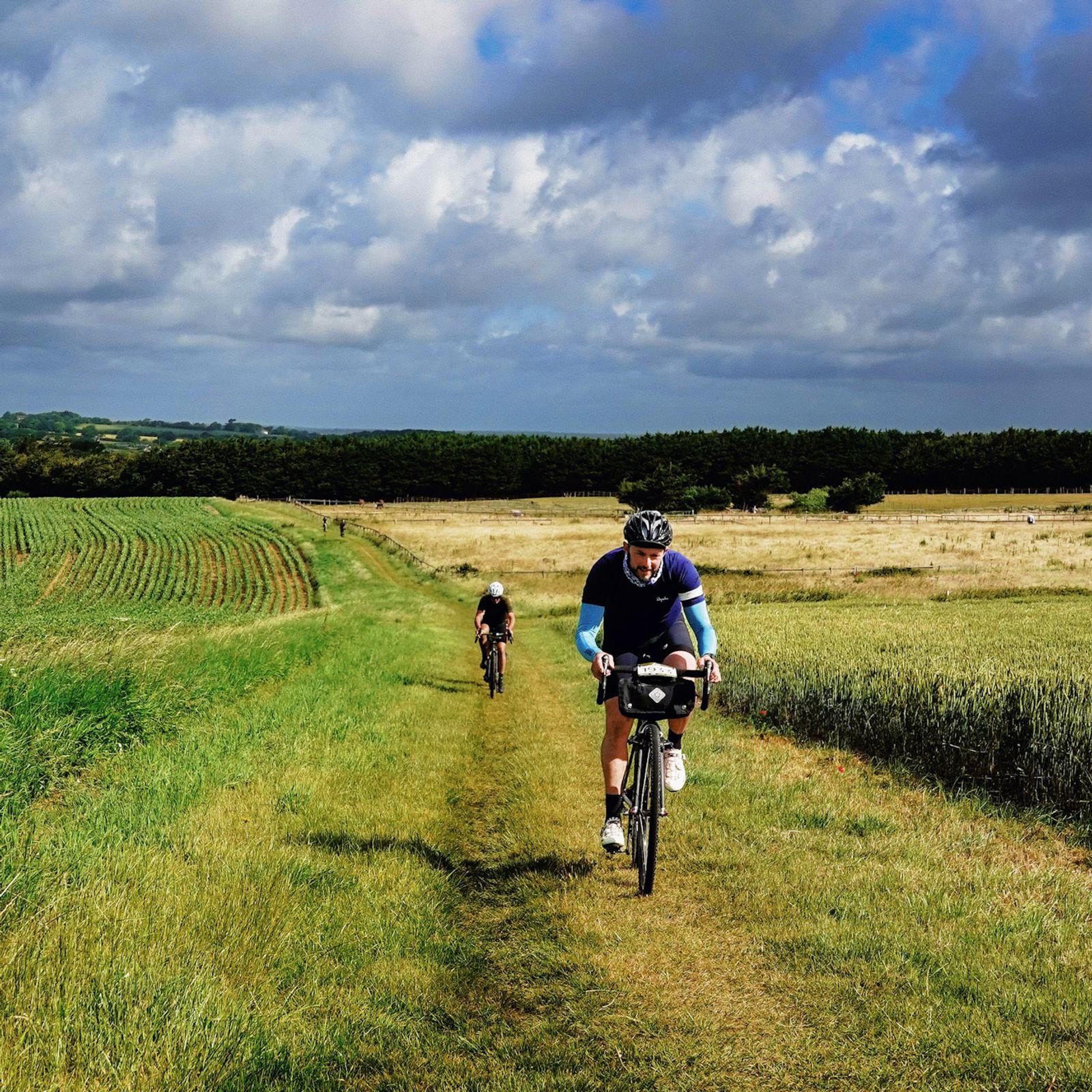 Two cyclists ride on a grassy trail through a rural landscape featuring fields and distant hills. The lead cyclist wears a blue and black outfit, followed closely by another cyclist. The sky is partly cloudy, casting a mix of light and shadows on the scene.