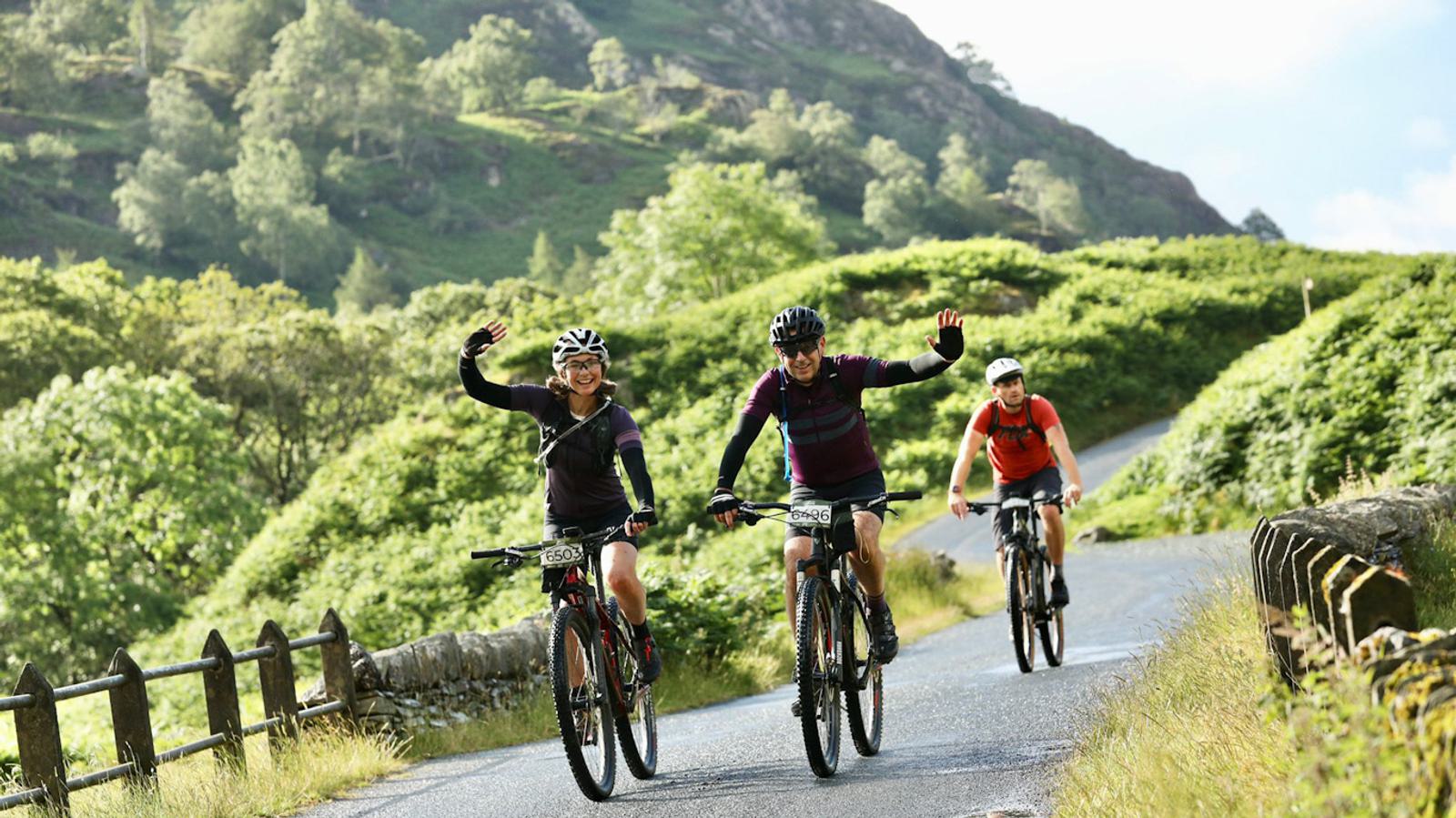 Three cyclists ride on a scenic, winding road surrounded by lush green hills and trees. Two of the cyclists in the front wave cheerfully, while the third cyclist follows behind, all wearing helmets and biking gear. Sunlight filters through the landscape.