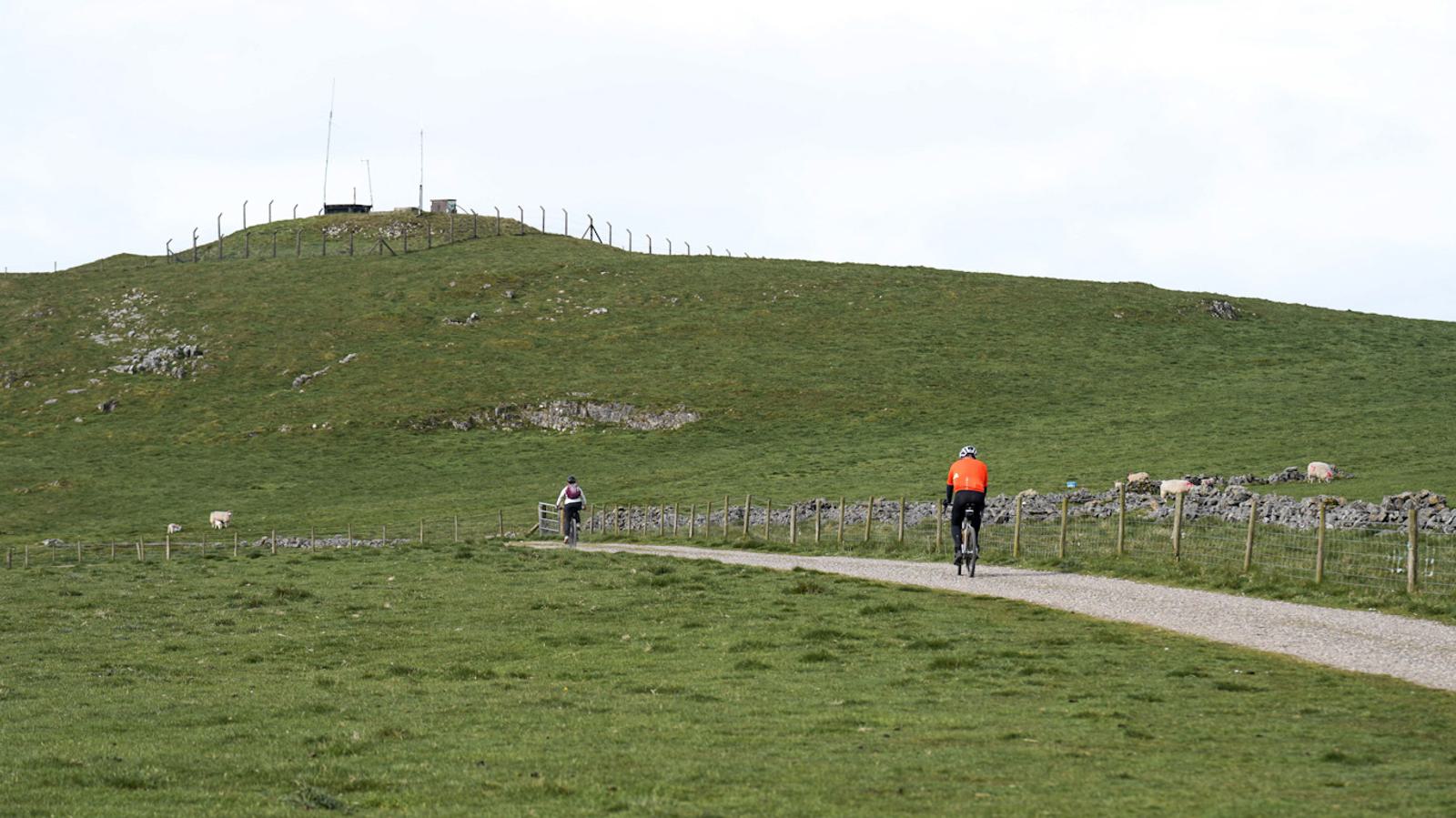 Two cyclists ride on a paved path through a grassy hillside. One cyclist wears an orange jacket, while the other is farther ahead, wearing a pink and white jacket. A small hill with antennas and a fence is visible in the background.