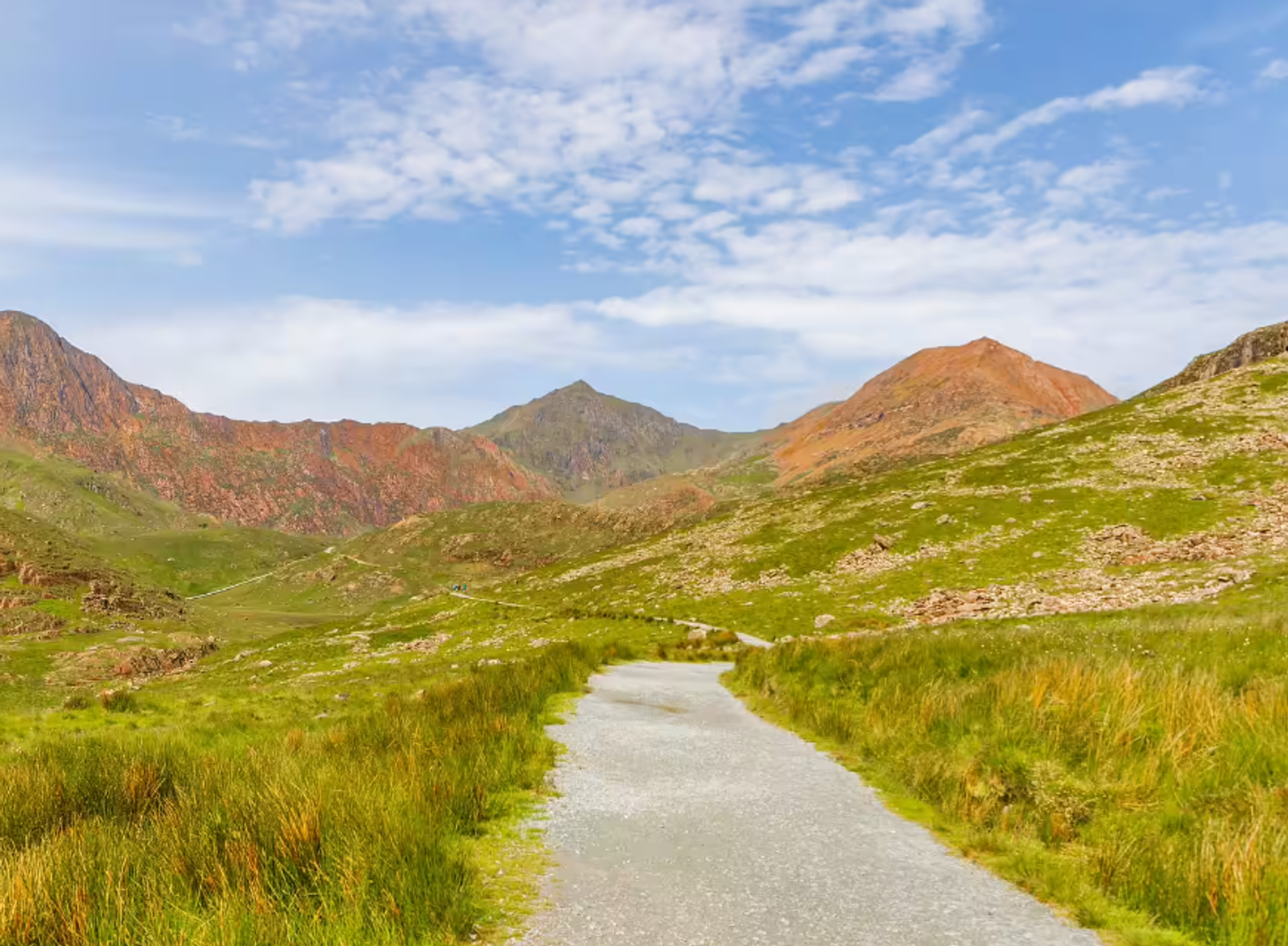 A scenic view of a gravel path leading through a grassy landscape with rolling hills in the distance. The hills are a mix of green and rocky terrain, under a partly cloudy blue sky.